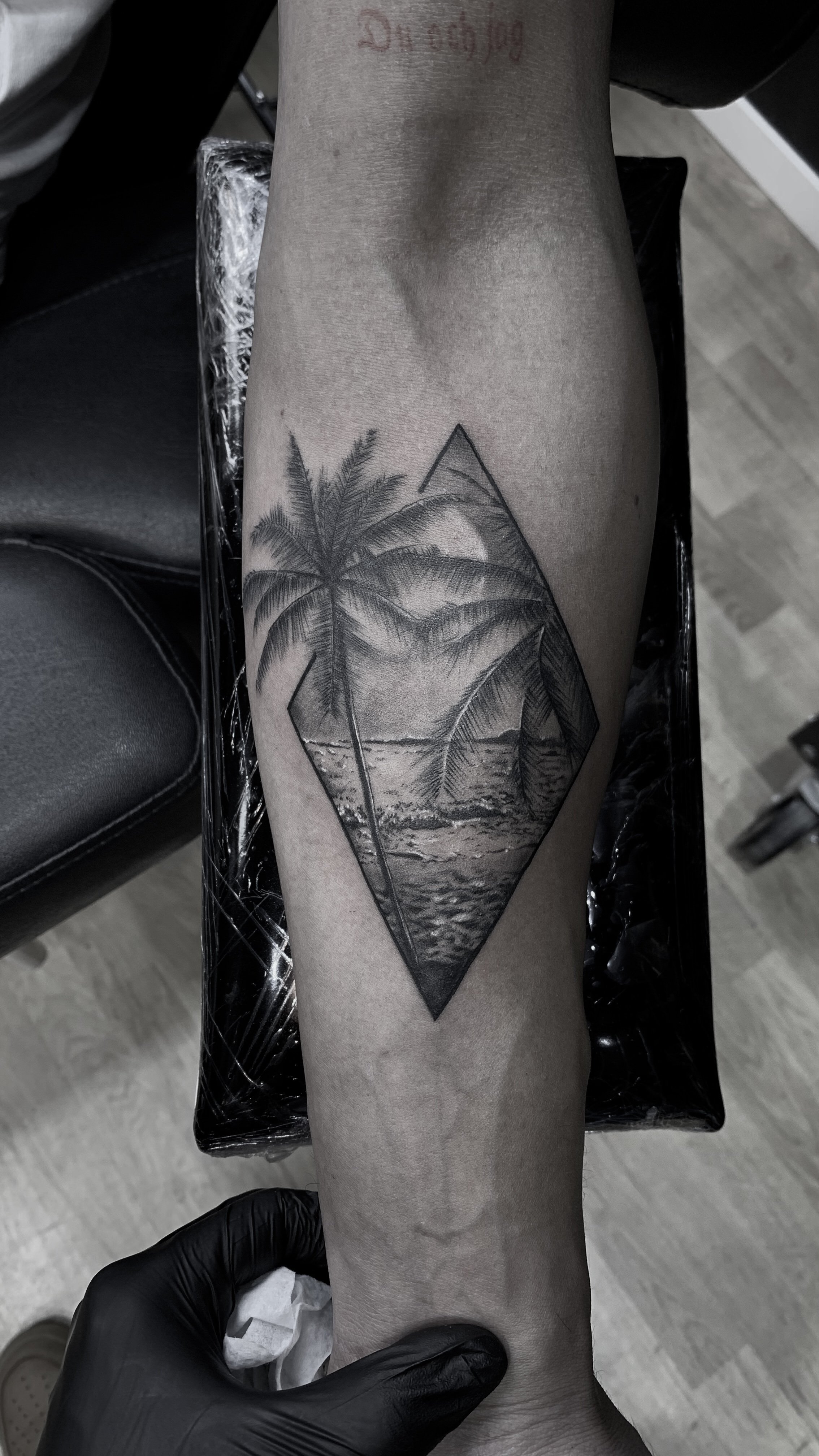 En tatuering på en underarm som föreställer ett landskap med palmer och en strand inom en diamantform.