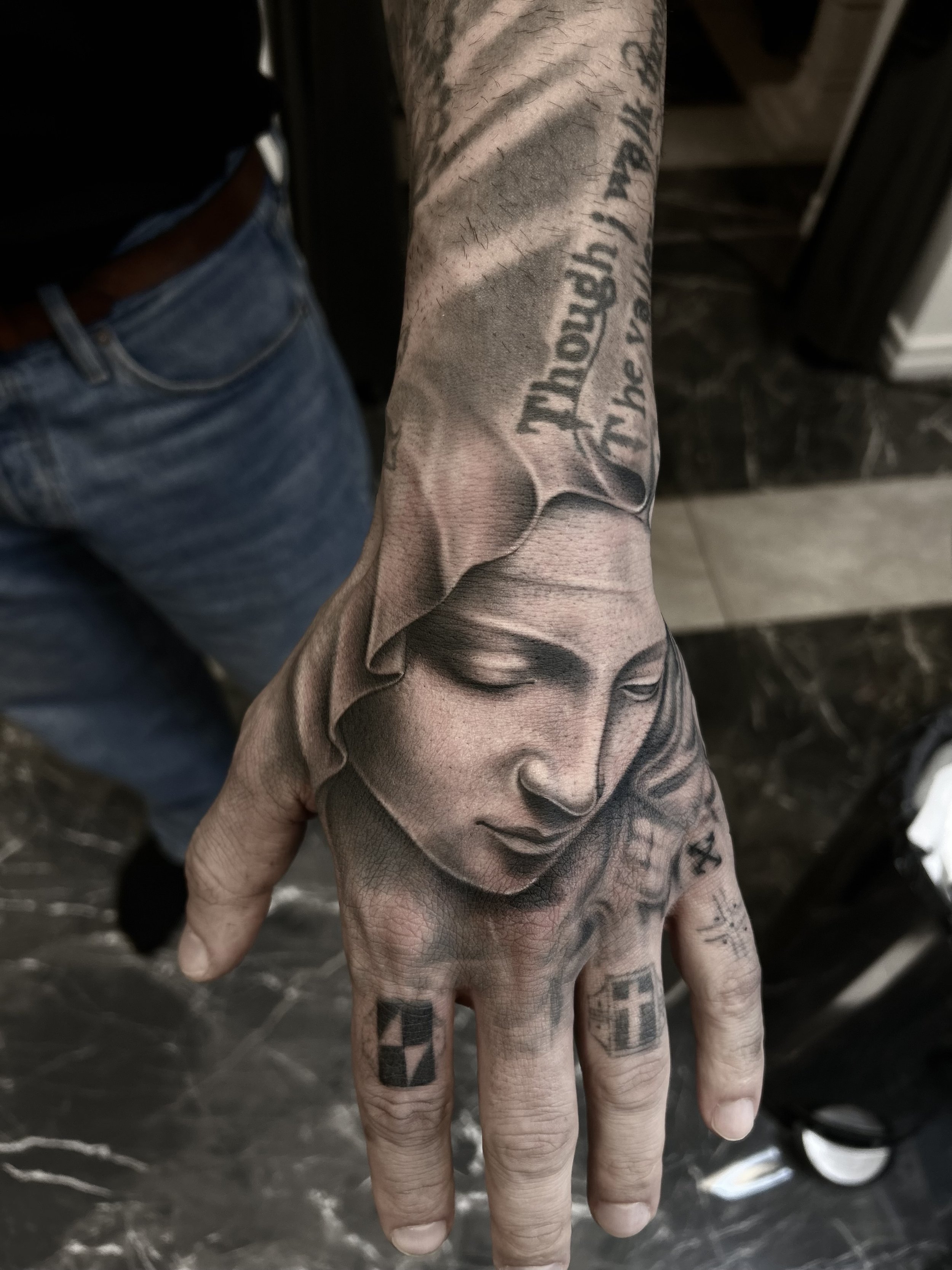 Hand med en tatuering av ett ansikte i ett klassiskt religiöst eller konstnärligt motiv, med texter och symboler på fingrarna.