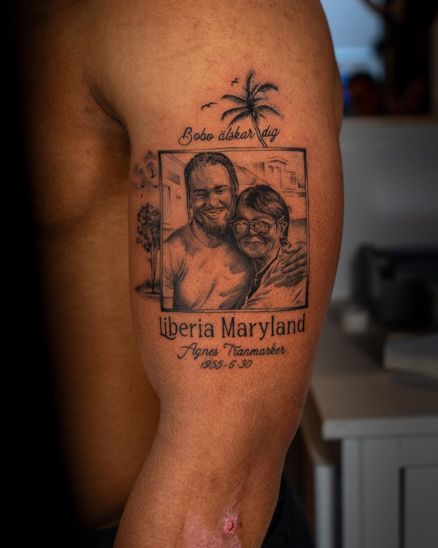 Tattoo on a person's upper arm featuring a portrait of two people inside a rectangle, with a small palm tree and birds above. The text includes 'Bobo älskar dig' and 'Liberia Maryland Agnes Framarker 1956-6-30.' The tattoo uses black ink.