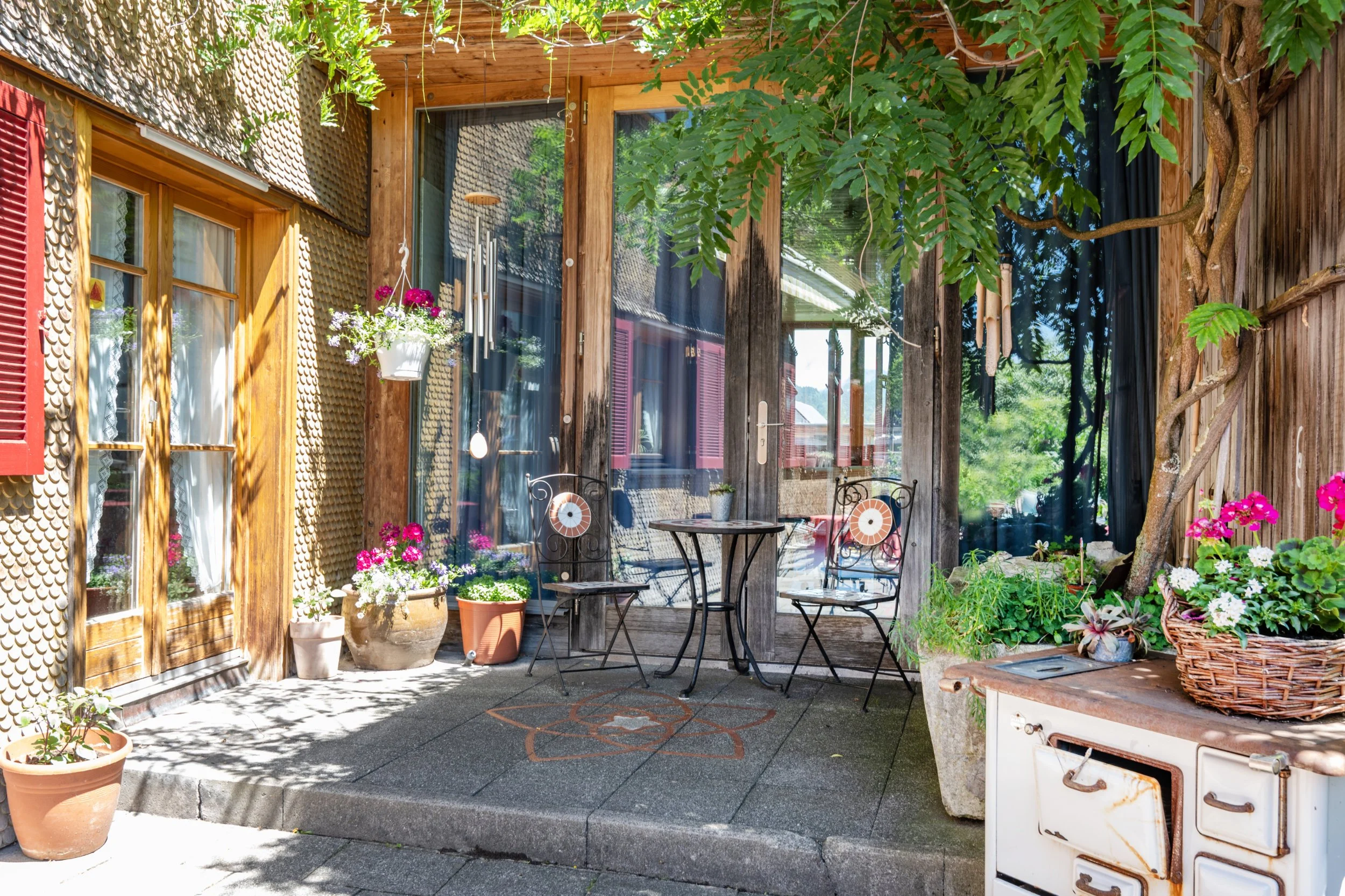 Gemütlicher Balkon mit Holzwand, bunten Blumen in Töpfen, Tisch und Stühle. Überdacht mit grünem Baumdach.