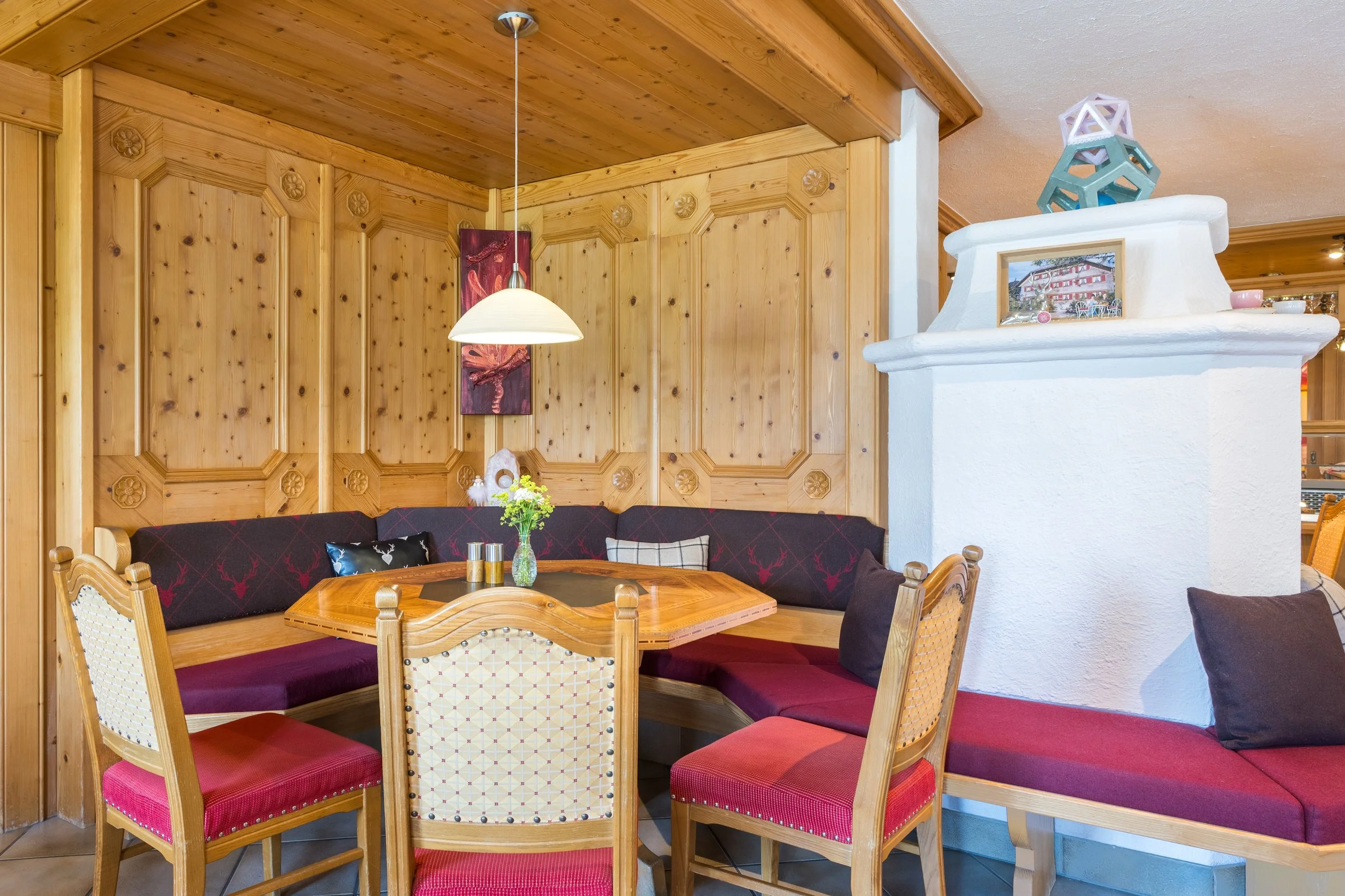 Ein gemütlicher Essbereich mit Holzmöbeln und roten Sitzkissen, Holzpaneelen an der Wand, eine weiße Kaminecke mit dekorativen Gegenständen und einem kleinen Familienfoto, eine Deckenlampe und ein kleiner Blumenstrauß auf dem Tisch.