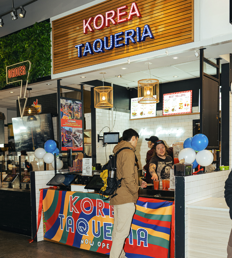 People ordering food at the Korea Taqueria counter in a shopping mall, decorated with balloons and a colorful banner.