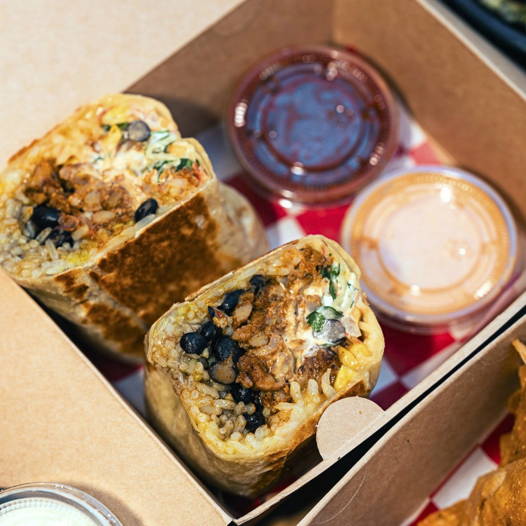 Two burritos cut in half, showing rice, beans, meat, and vegetables inside, with small containers of salsa and sauce on a red and white checkered paper in a cardboard takeout box.