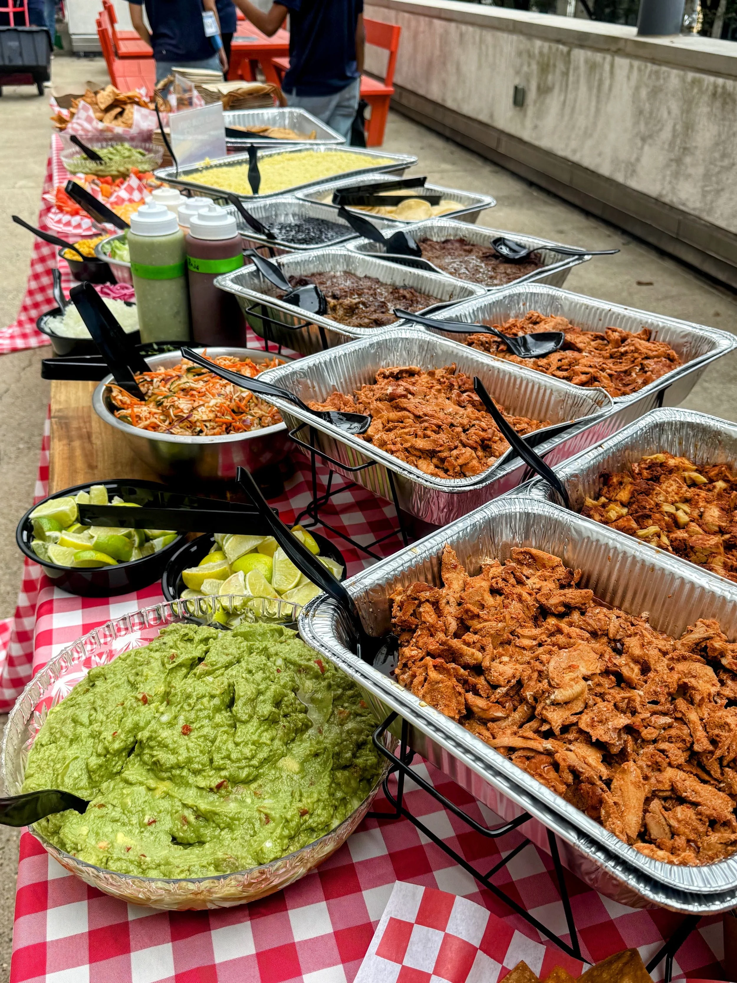A buffet-style spread of Mexican food on a checkered red and white tablecloth, including trays of shredded meat, beans, rice, guacamole, lime wedges, coleslaw, and condiments.