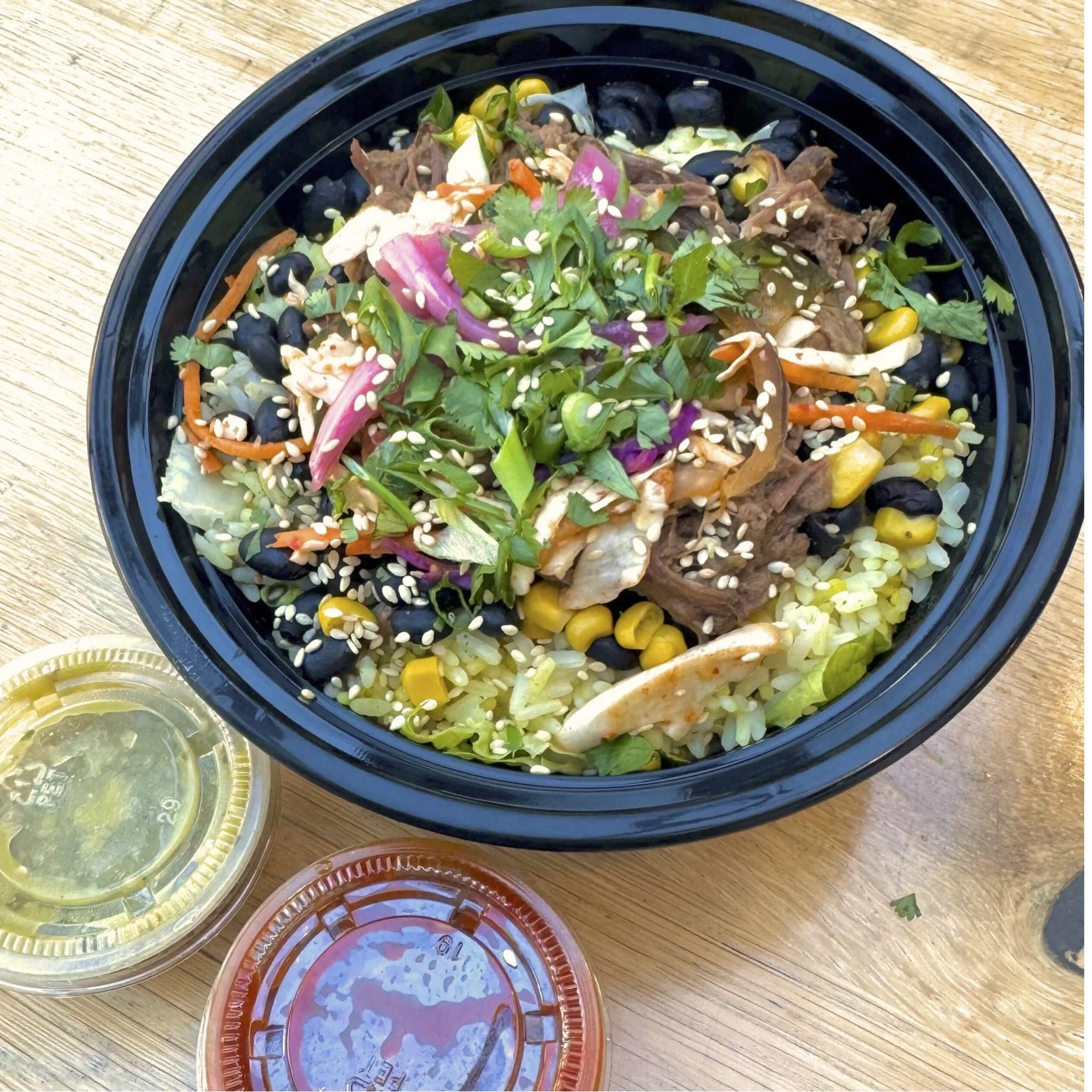 Large bowl of Asian-style salad with shredded beef, rice, corn, black beans, sliced vegetables, cilantro, sesame seeds, and sauces on the side on a wooden table.