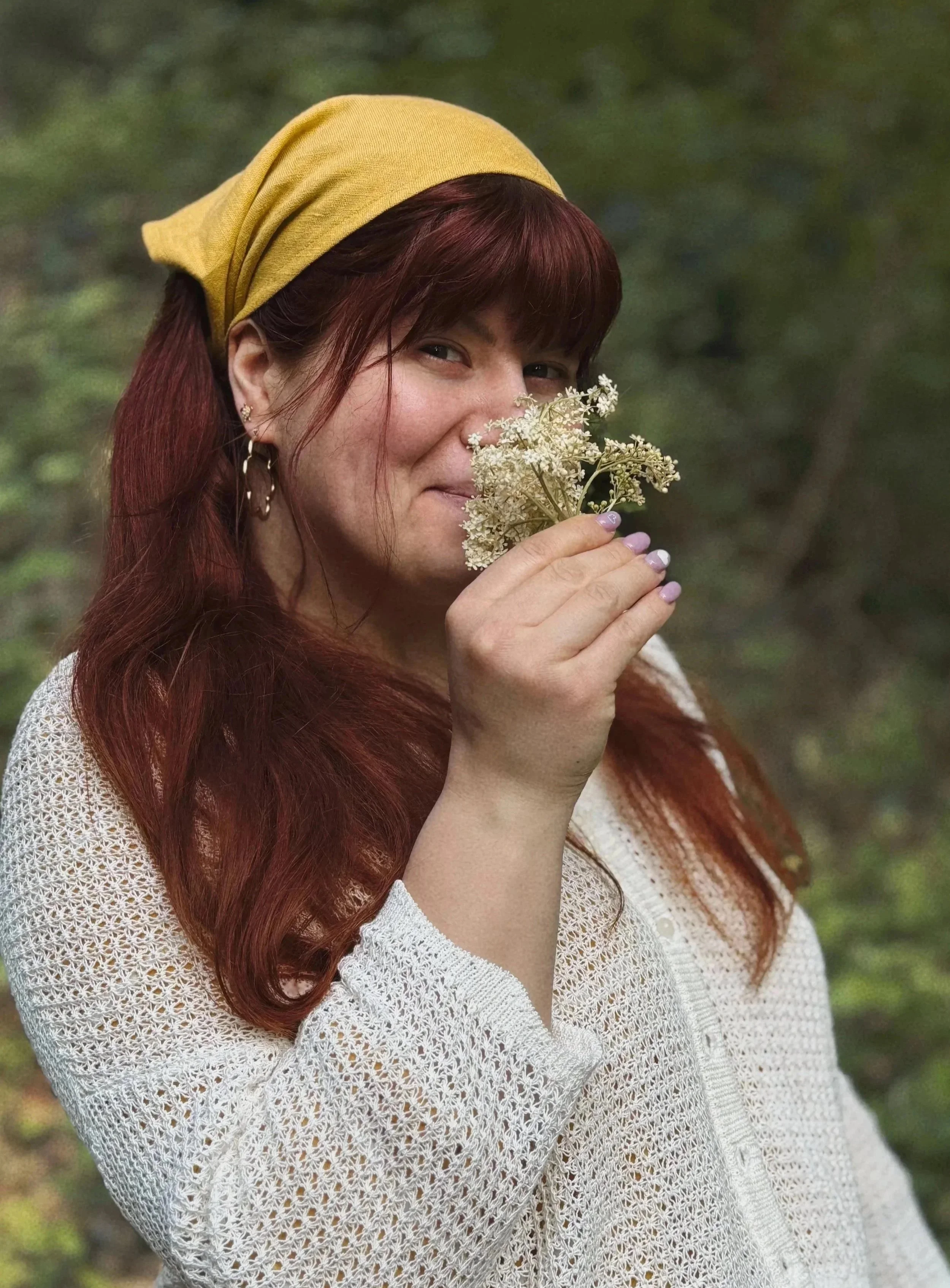 Eine Frau mit roten Haaren, gelber Kopfbedeckung, weißen Strickpullover, hält eine weiße Blumen in der Hand und lächelt leicht.
