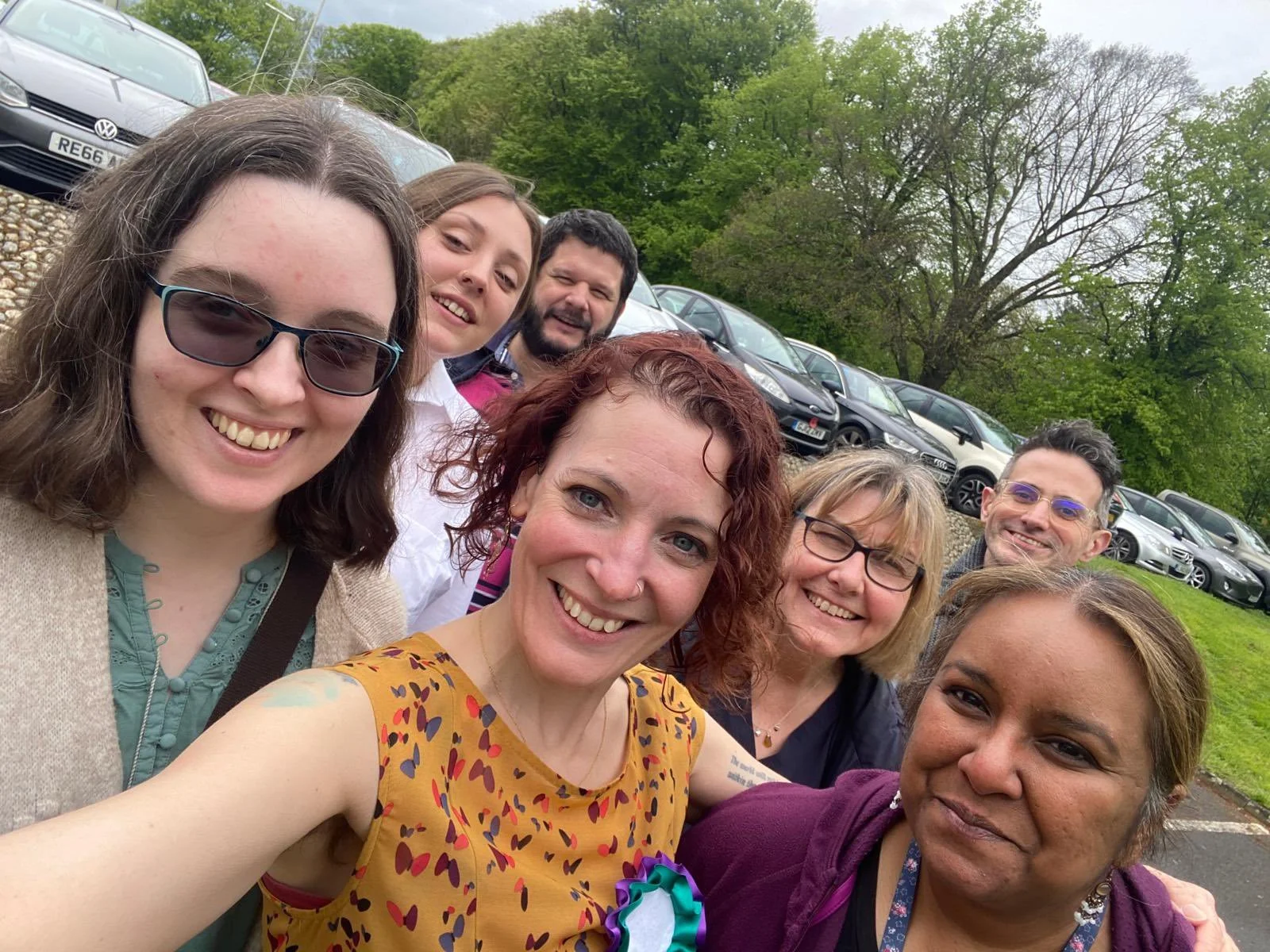 A group of seven people smiling outdoors, with a row of cars and trees in the background. The setting appears casual and relaxed.