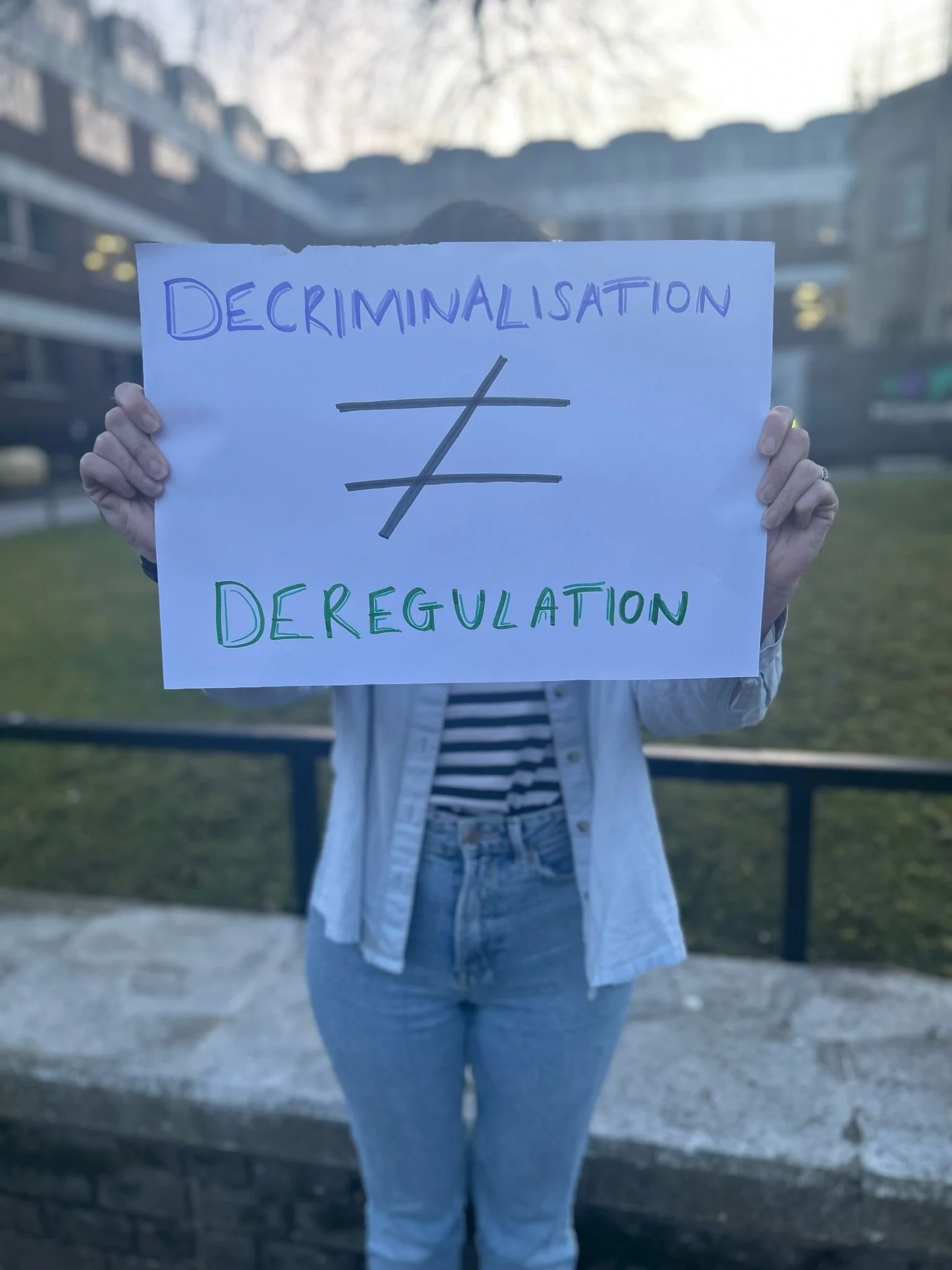 Person holding a sign reading "Decriminalisation ≠ Deregulation" standing outdoors in front of the Basingstoke & Deane Borough Council building.