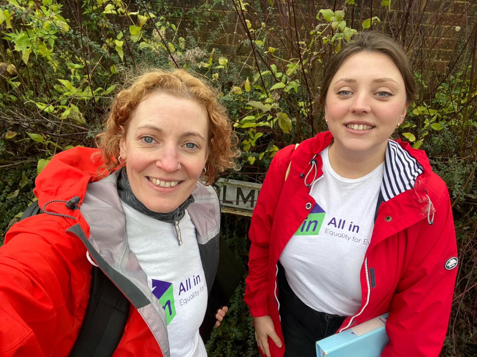 Two women smiling at the camera. One has red curly hair and one has brown hair tied back, but they're both wearing All In Party branded t-shirts and red waterproof coats