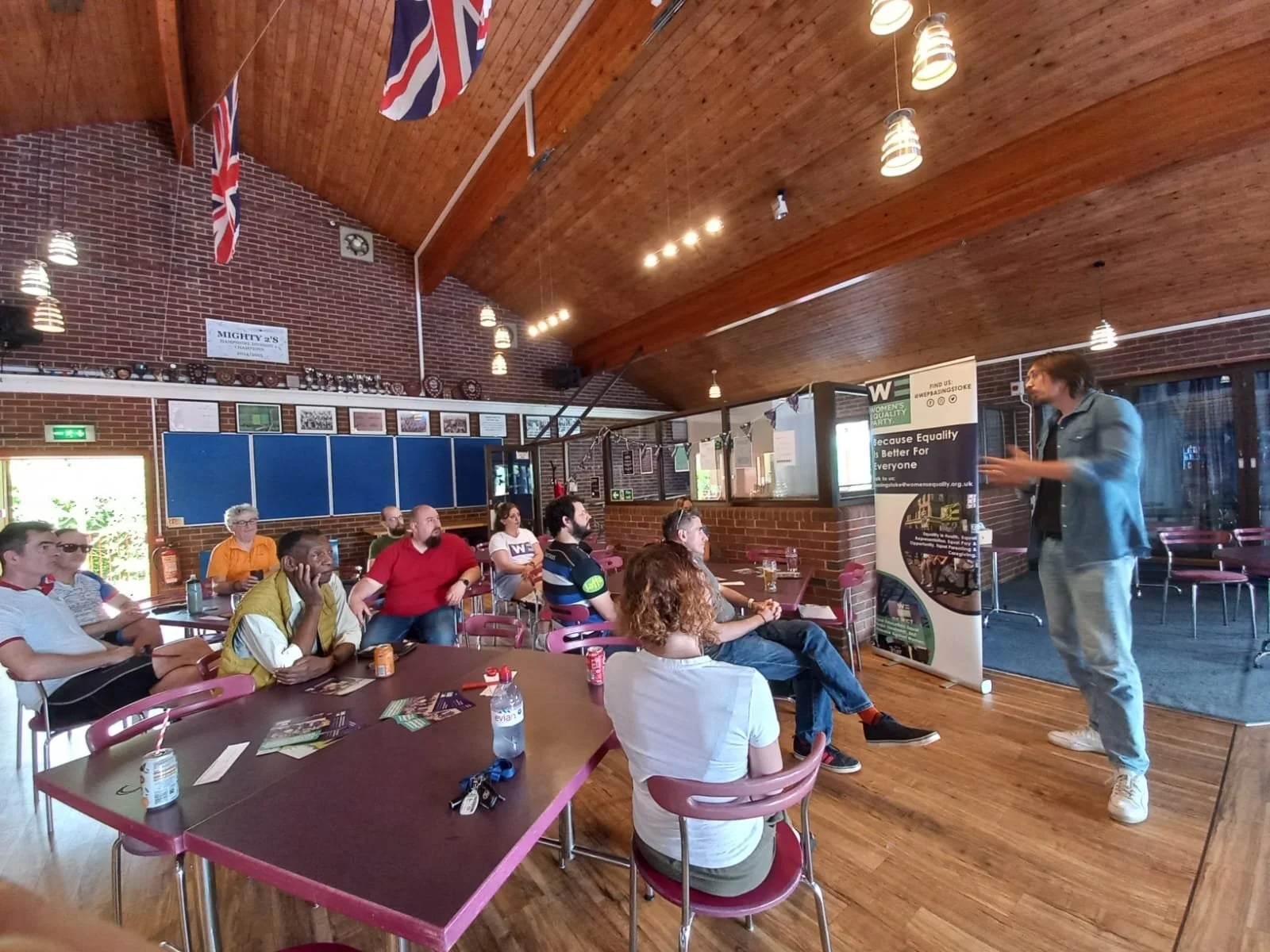 A group of men sitting at tables listening to a male speaker in a room with brick walls. The speaker is standing next to a banner. The room has wooden floors and a high ceiling.