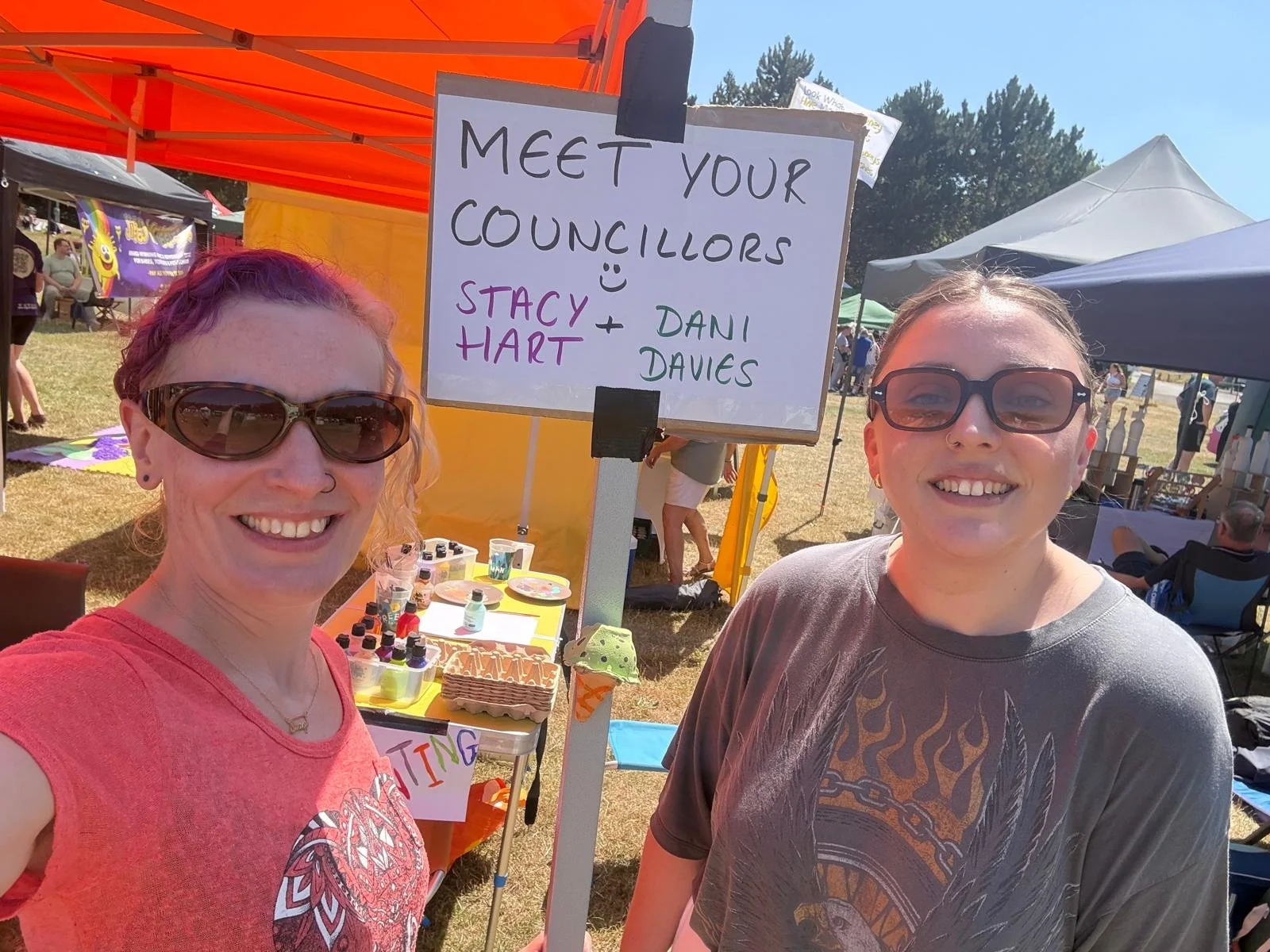 Two people in a field standing next to a sign, encouraging people to ask them a question about our work