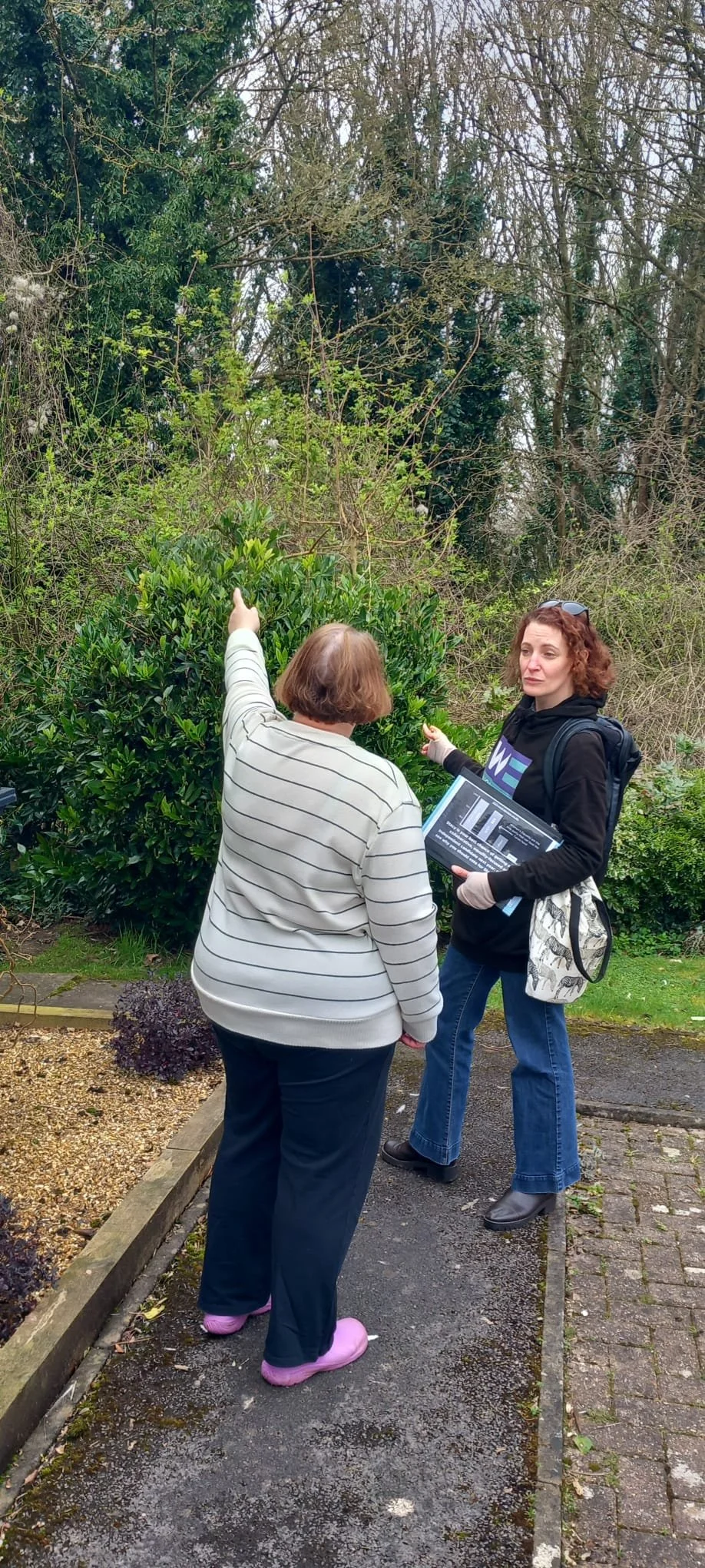 Two women in a garden, pointing at trees and bushes, one holding a sheet of paper.