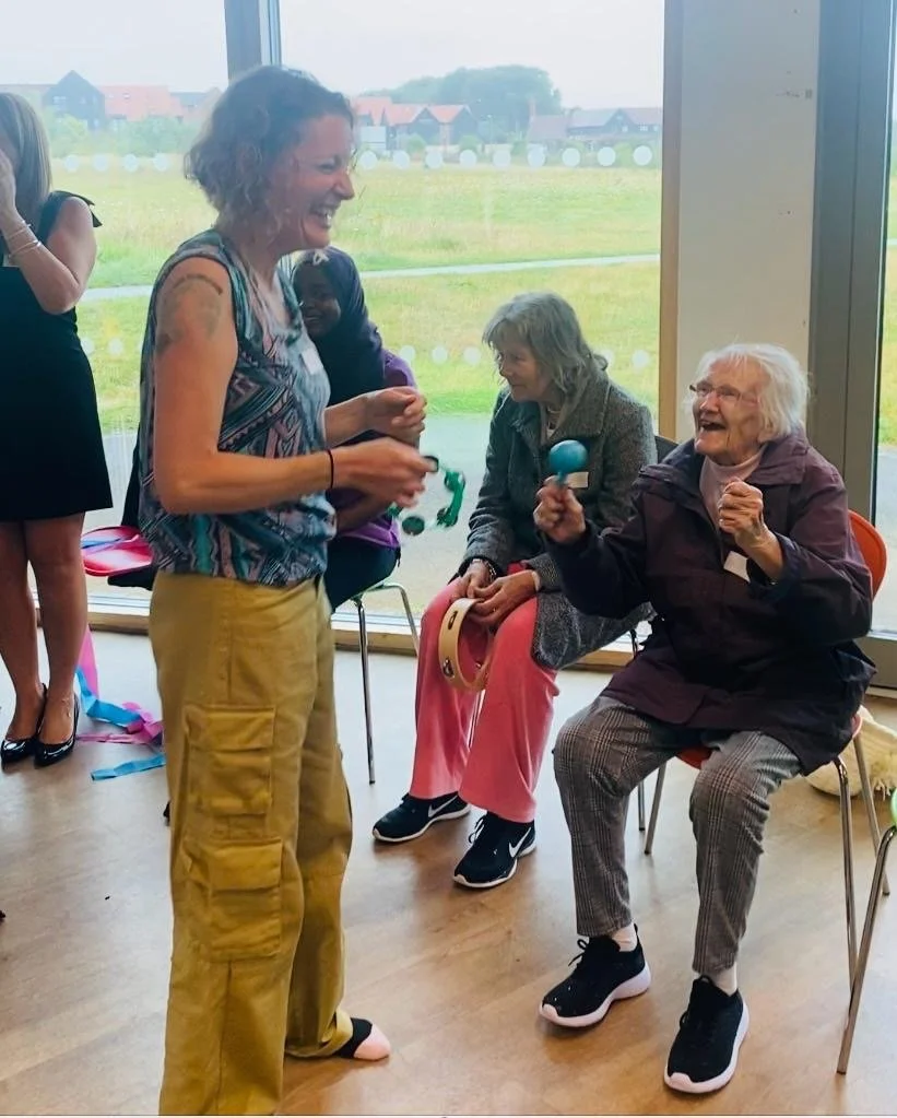Group of people participating in a music activity, some seated, using percussion instruments like maracas and tambourines, with a woman standing and smiling. Large windows show outdoor view in background.