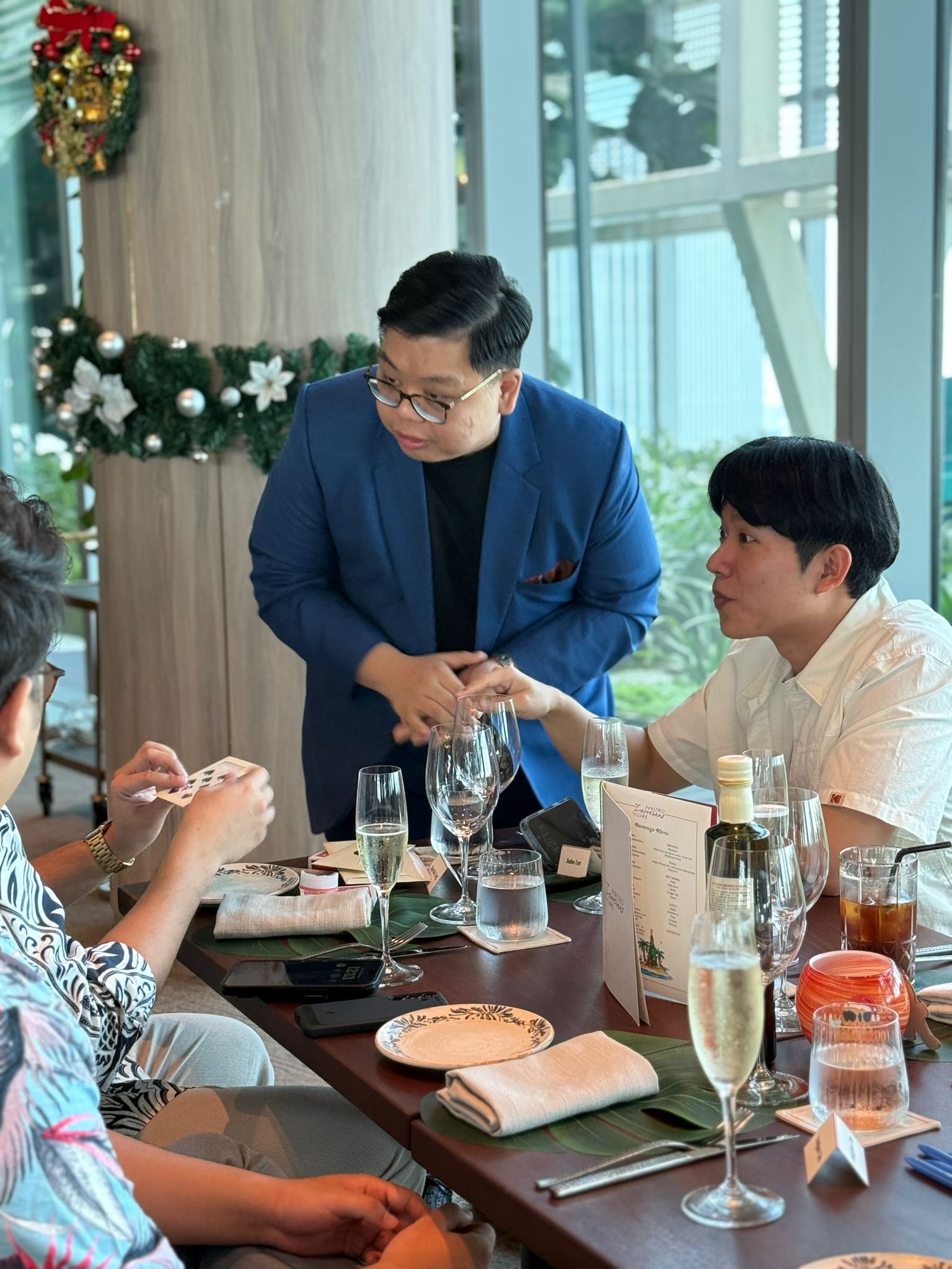 People at a restaurant table during a celebration, with decorated Christmas wreaths and ornaments on the wall, and a man in a blue blazer shaking hands with a seated man in a white shirt.
