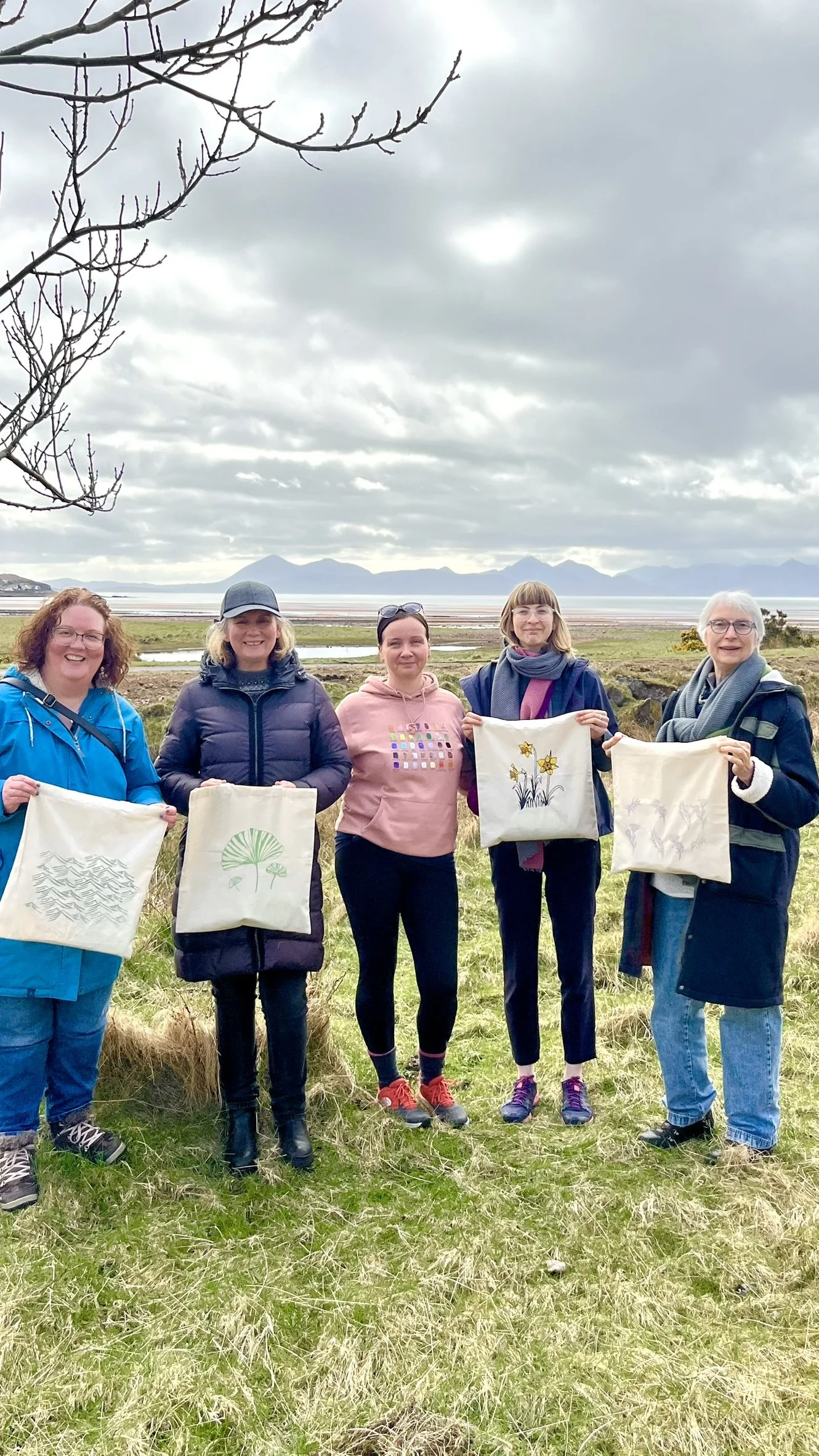 Five women standing outdoors on a grassy field holding tote bags with designs they have handprinted during a workshop with Tery, with mountains and cloudy sky in the background.
