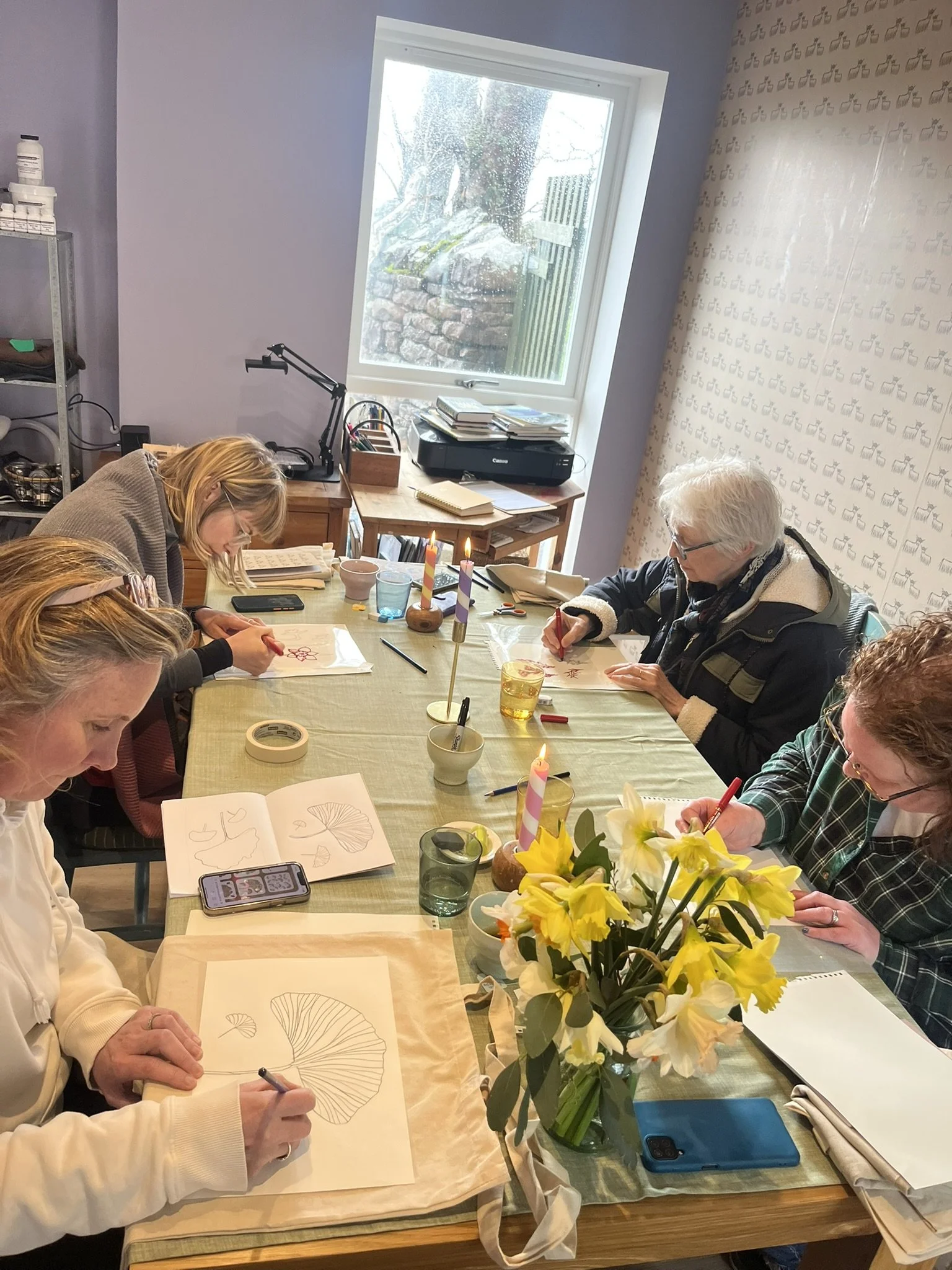 A group of four women sitting around a table engaged in drawing or art activities, with flower arrangements and lit candles in the center of the table, in a room with a window showing outdoor rocks and trees.