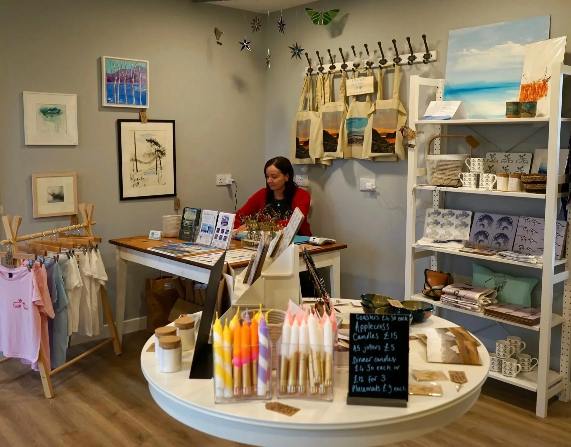 Applecross Interiors and Gifts shop with art prints, home decor, clothing, and candles for sale. There are handmade tote bags on the wall, and a shelf with mugs, art prints, and woven baskets.
