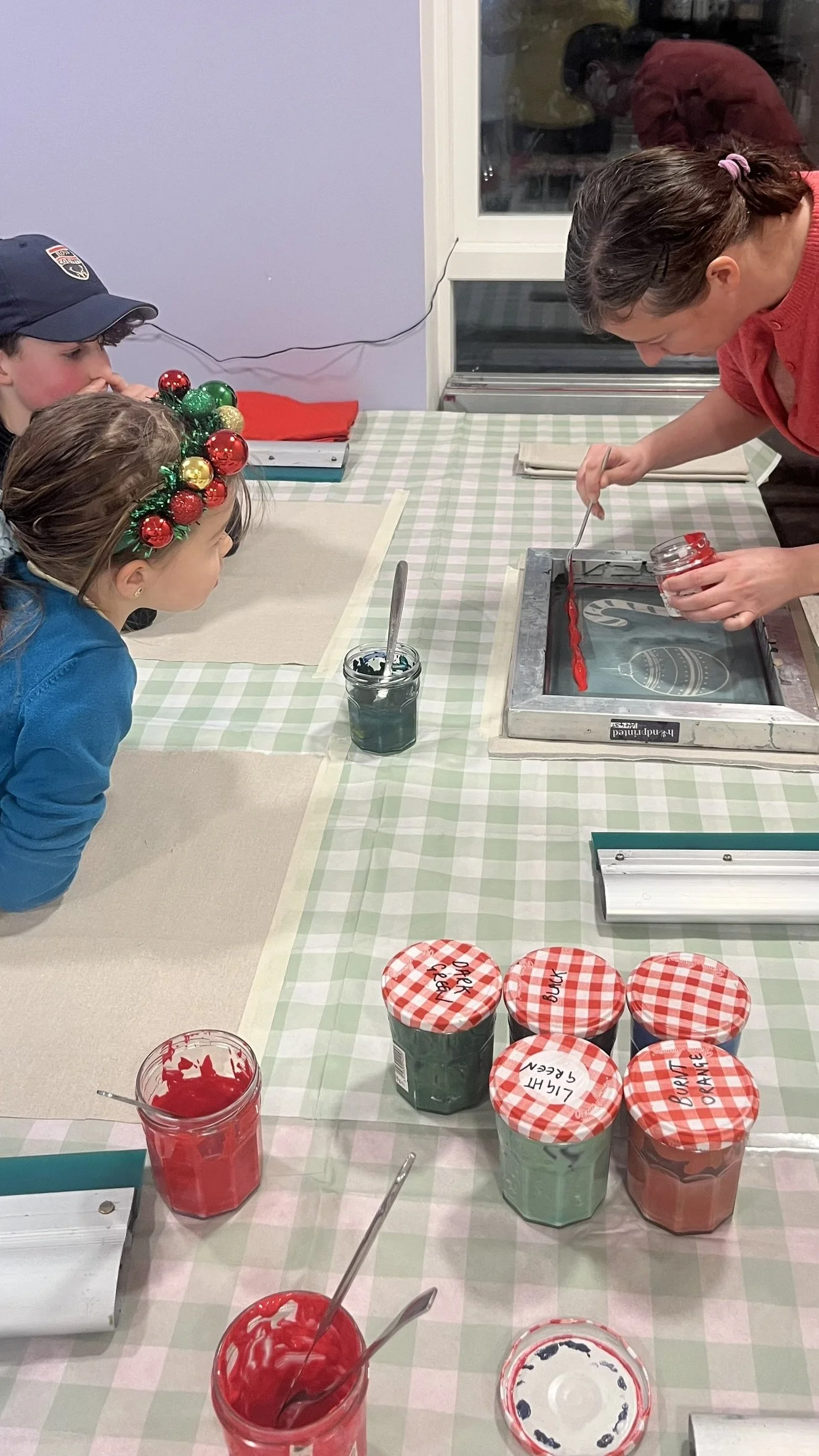 A screen printing workshop where the table is covered with a green and white checkered tablecloth, and there are jars of red and green paint.