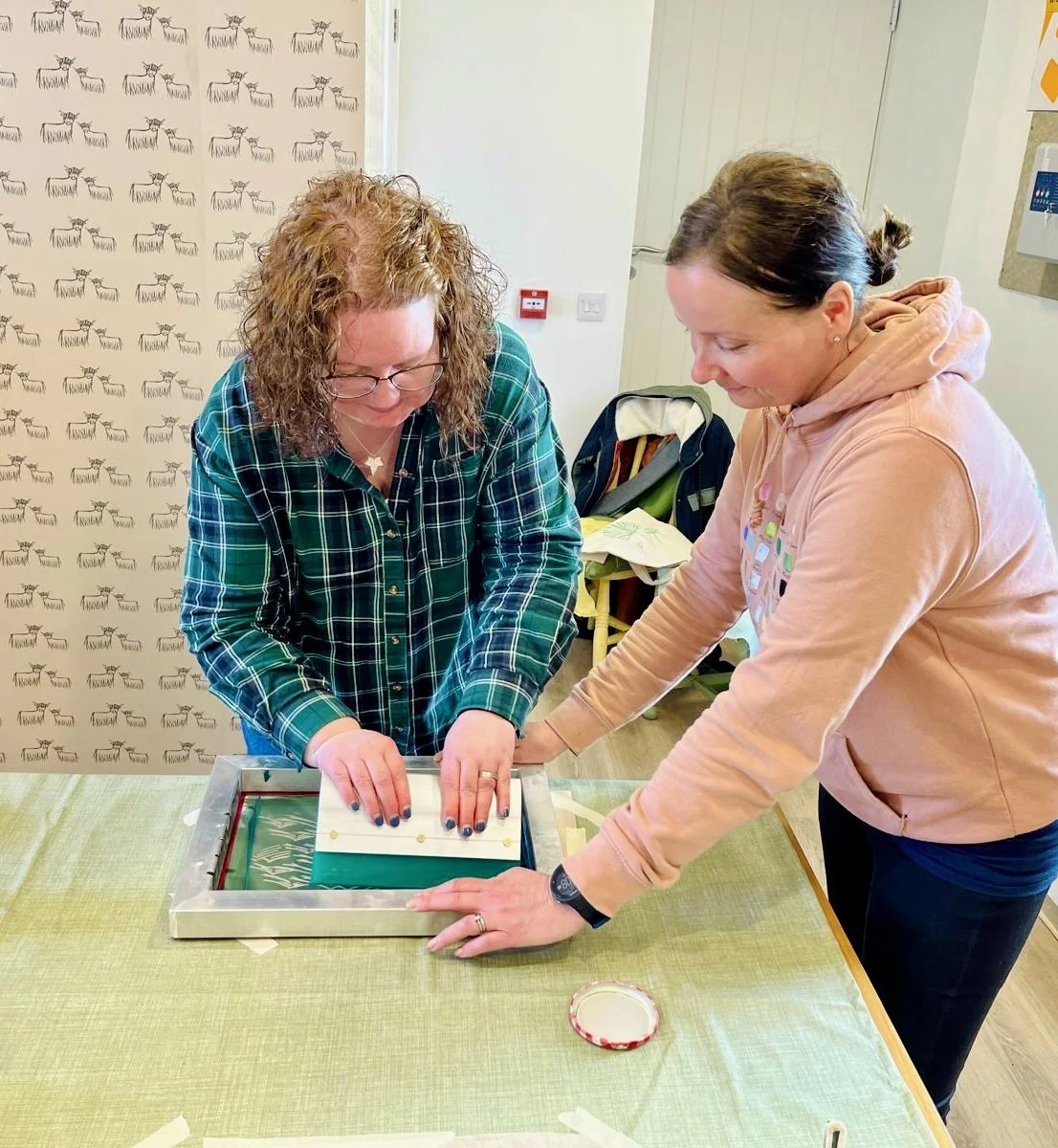 Two women are working together on a craft project at a table. One is pressing down on a piece of fabric over a frame, while the other supports with her hands. Behind them is a wall featuring Clement Design deer pattern wallpaper.
