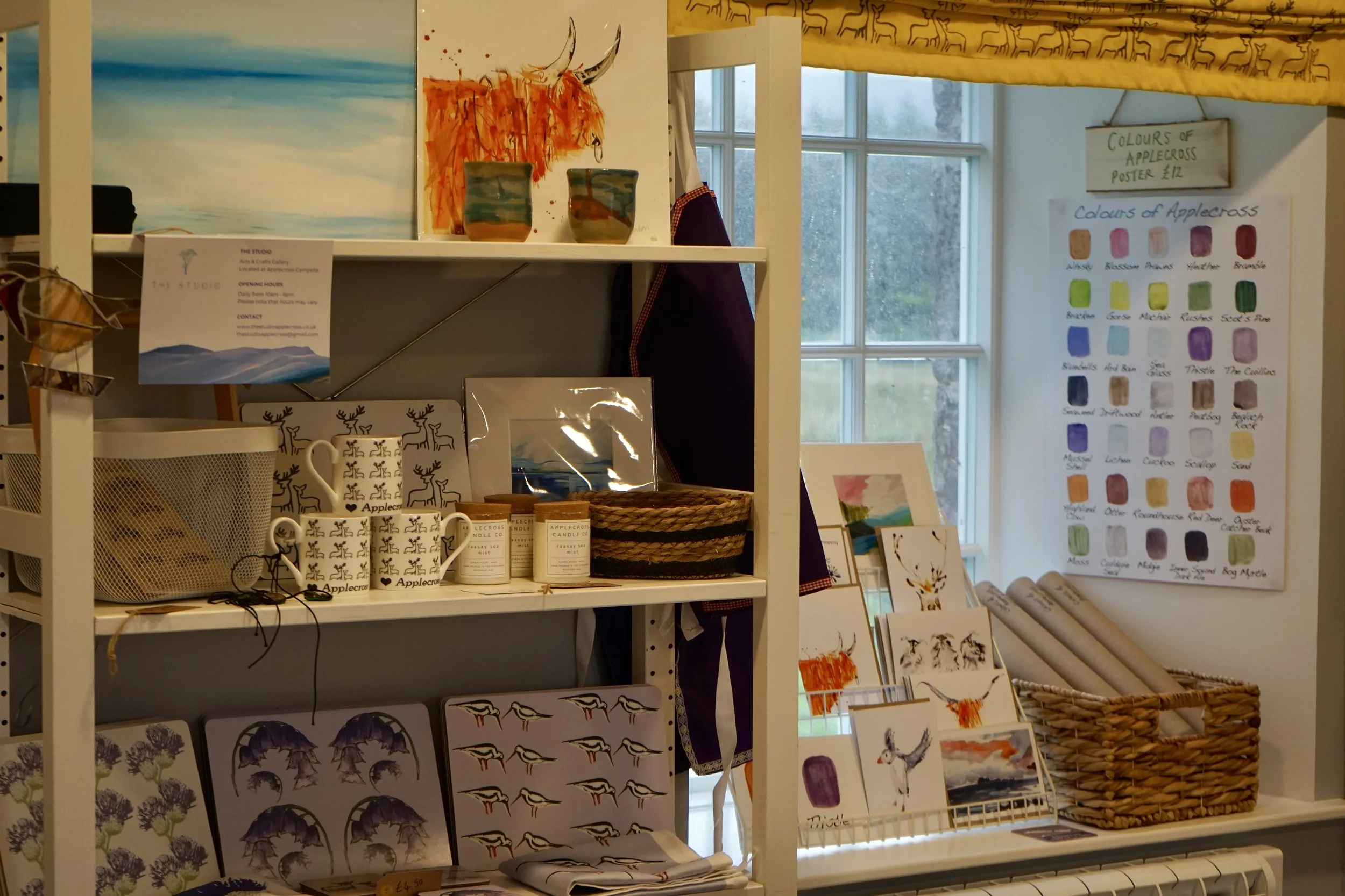Display shelf with art prints, mugs, candles, and decorative items in a shop, with a window in the background.