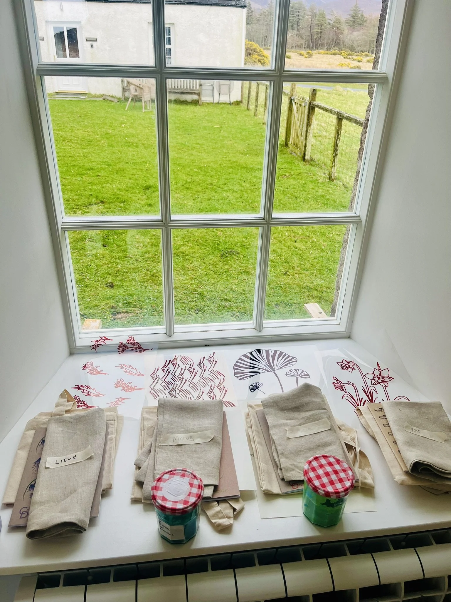A window overlooking a green backyard with a white house and a wooden fence. On the windowsill, there are folded cloth napkins labeled with names, two jars of paint with red checkered lids.