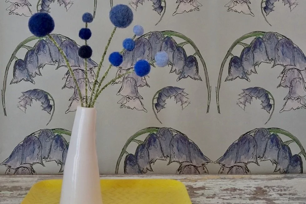 Decorative vase with blue and purple felted pom-poms on wire stems, positioned on a yellow tray in front of Clement Design bluebell print wallpaper.