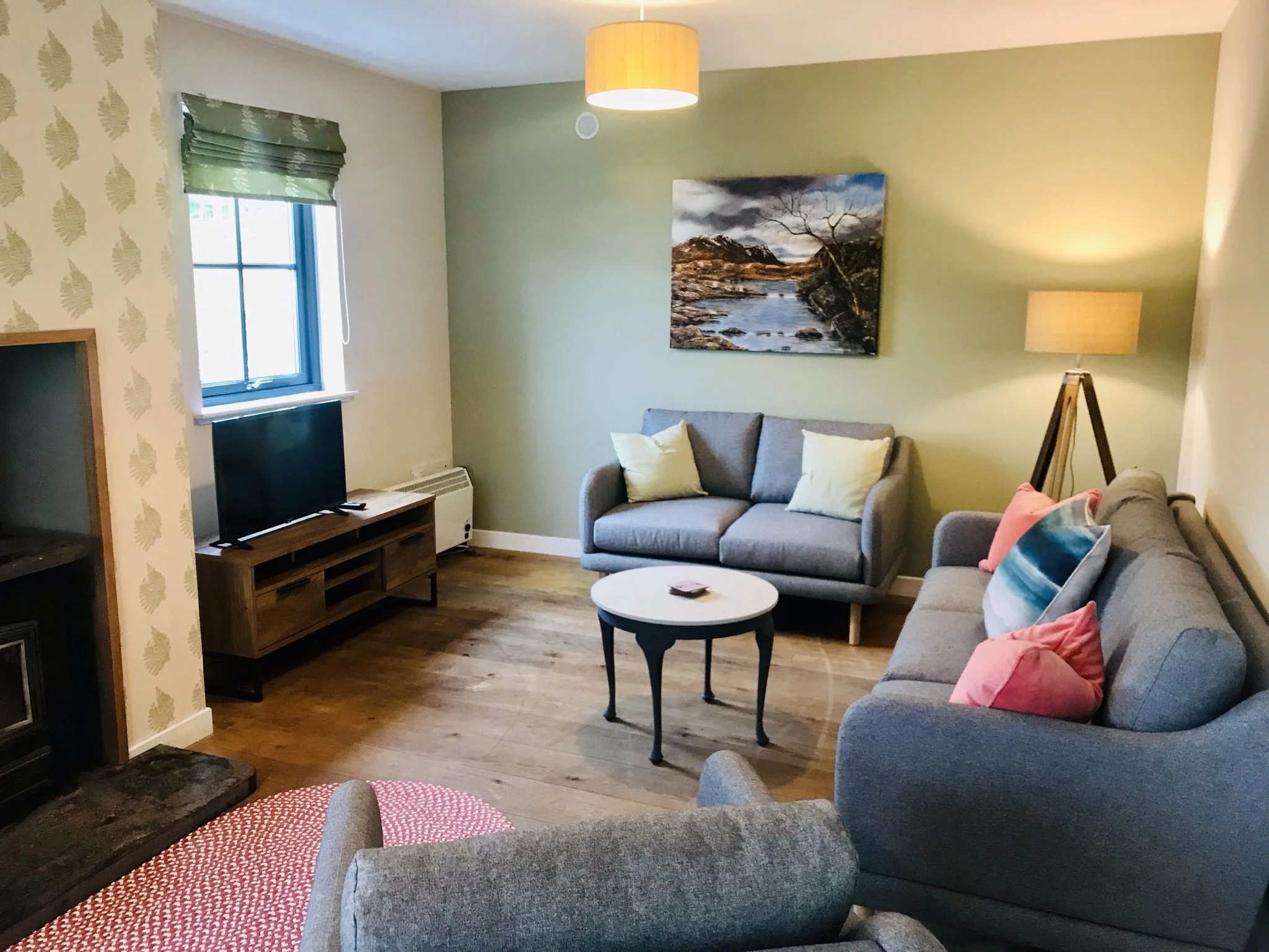 Living room with wallpaper and blinds by Clement Design, two gray couches, one with pink and blue pillows, a small round coffee table, a wooden TV stand with television, a painting of a river landscape, a standing lamp, and a patterned rug.