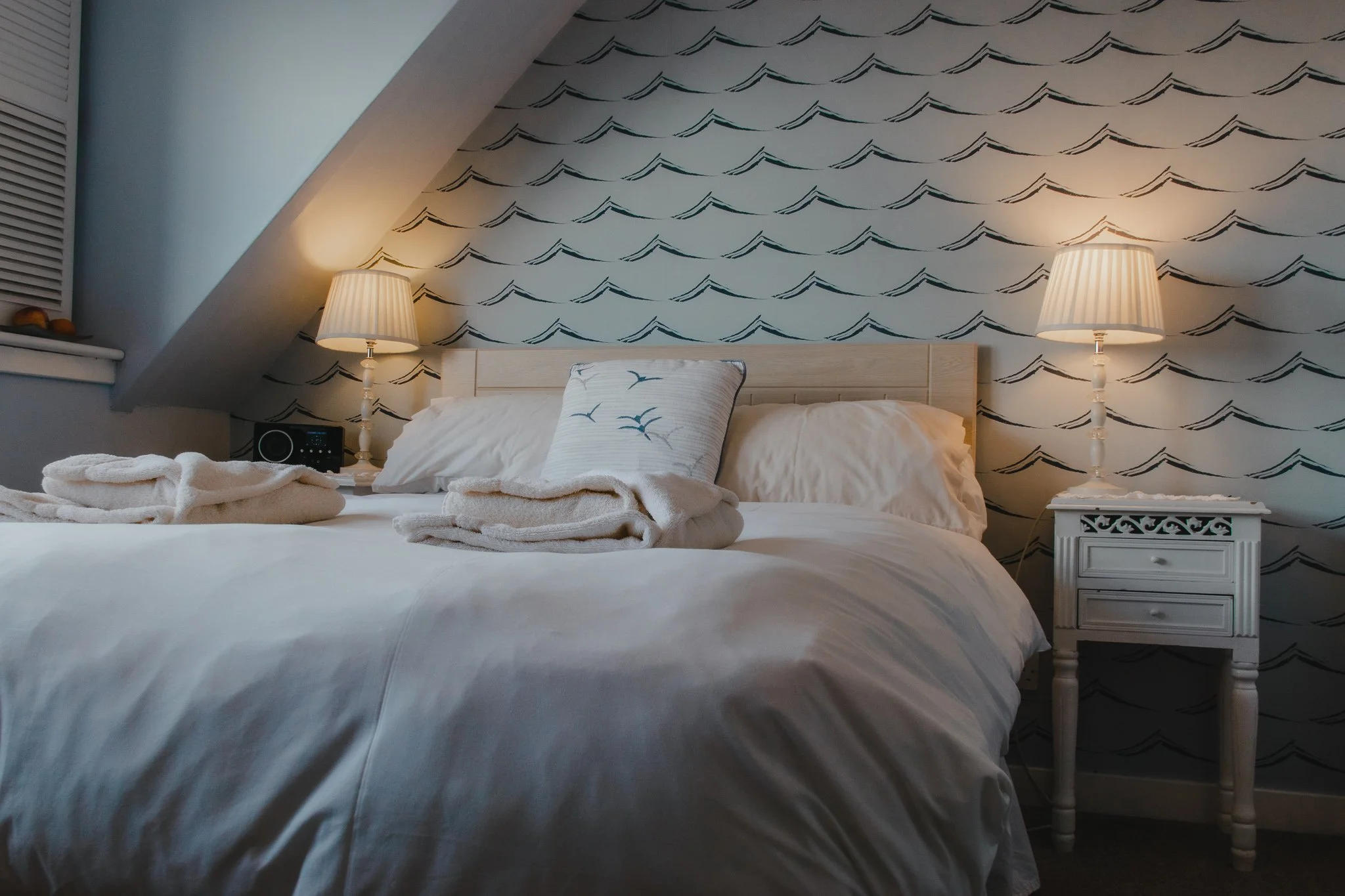 A cozy bedroom with a sloped ceiling, featuring a double bed dressed in white linens with a matching pillow and a decorative pillow with a seabird pattern. The wallpaper behind the bed showcases Clement Design's wave print wallpaper.