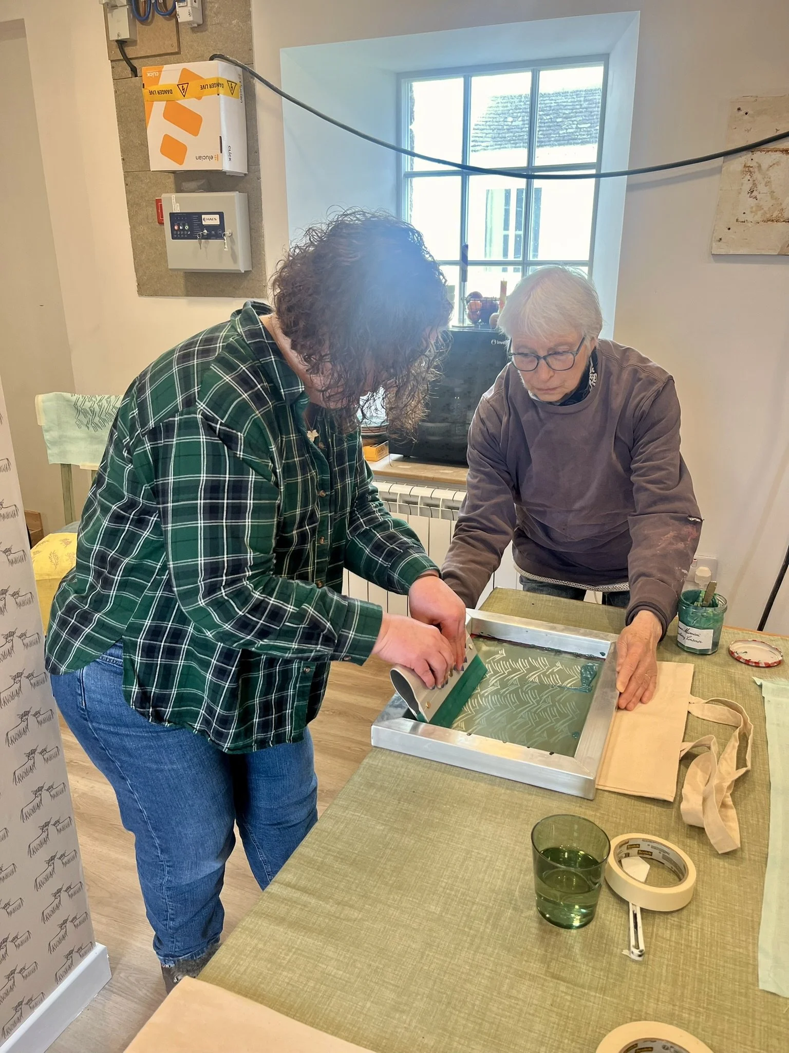 Two women working together on a craft project at a table in a well-lit room with a window in the background. One woman is wearing a green checkered shirt and the other is wearing a brown long-sleeve shirt. They are handling a screen printing frame.