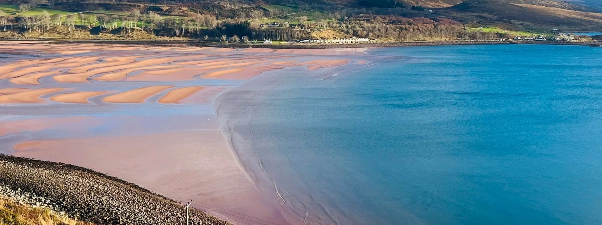 View from Applecross Interiors and Gifts overlooking shores Applecross Bay and blue ocean water on a sunny day, with hills and green fields in the background.