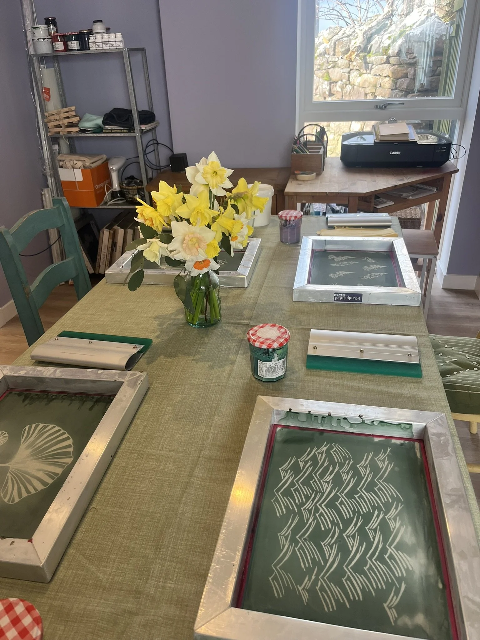 A craft table with screen printing panels, a vase of daffodils, jars, and other supplies, in a bright room with a large window.