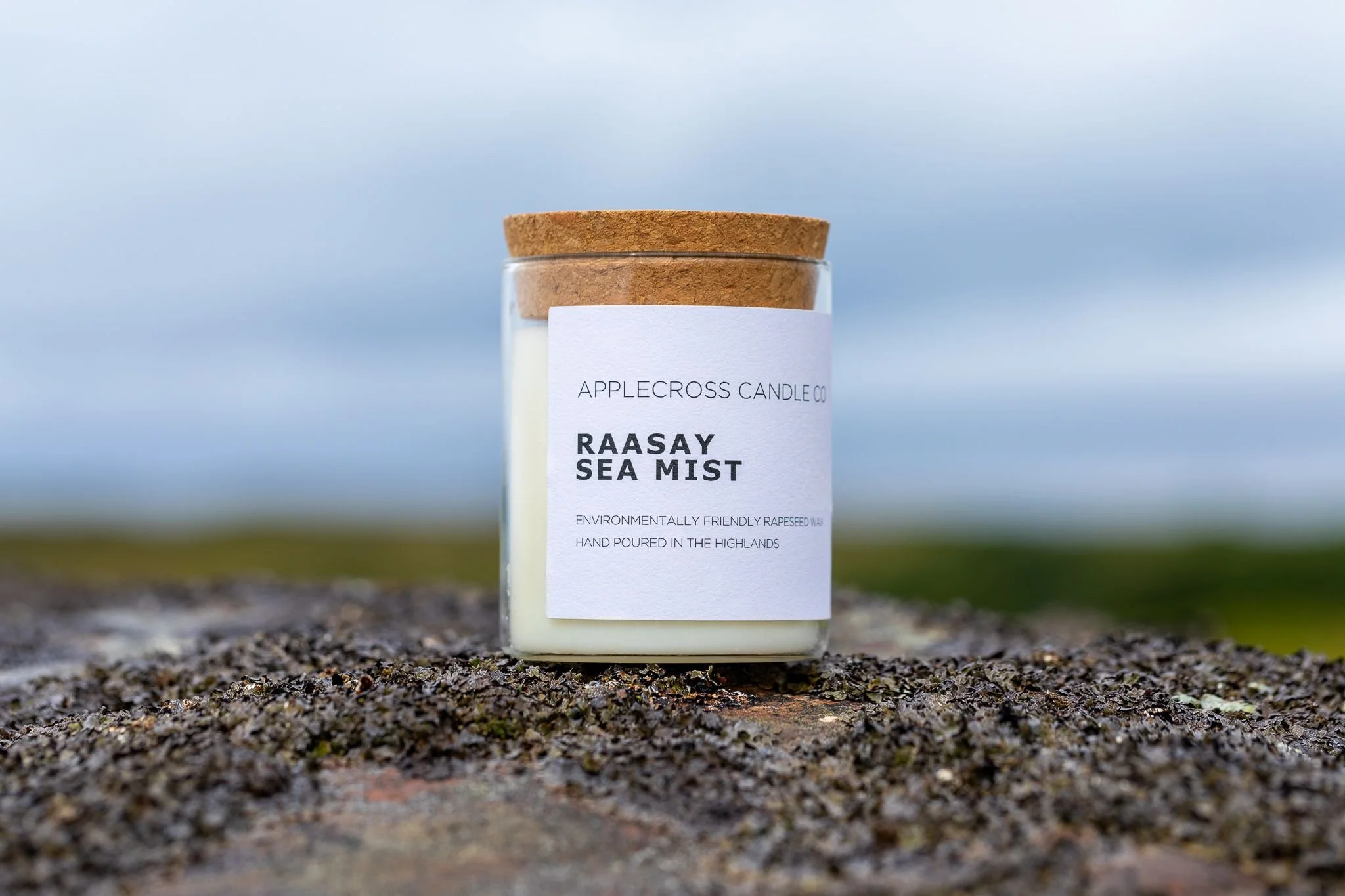 A white candle in a glass jar with a cork lid, labeled 'RAASAY SEA MIST' from Applecross Candle Co, placed outdoors on a textured surface with a blurred natural background.
