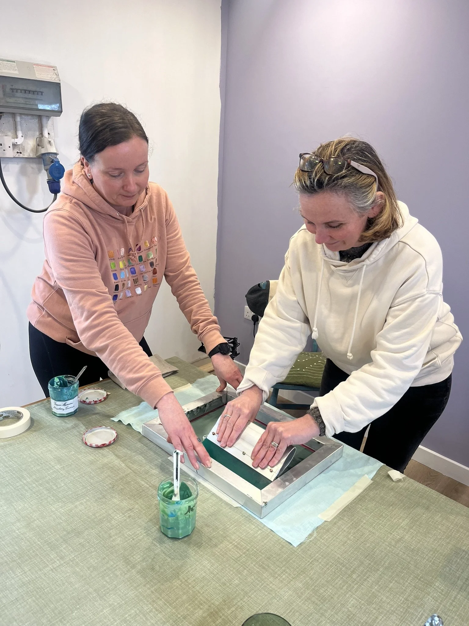 Two women are working together on a creative project at a table, using a screen printing frame with green ink in a room with a light purple wall.