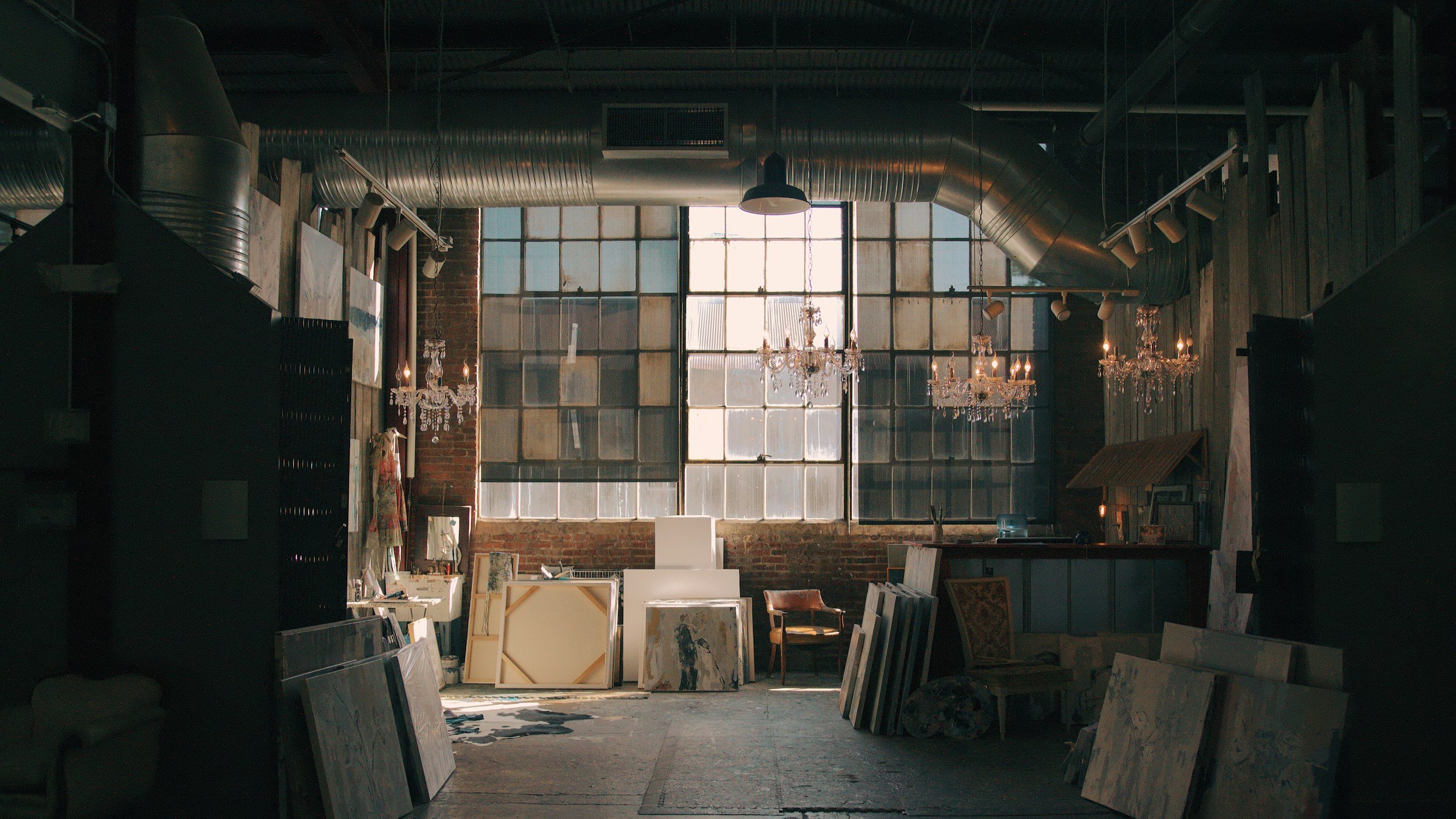 Interior of an art studio with large industrial windows, exposed ductwork, chandeliers, and various paintings and canvases leaning against the walls.