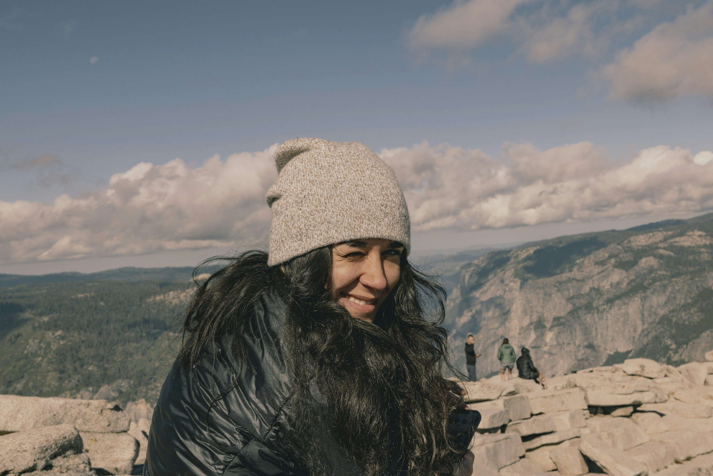 Woman with long dark hair wearing a gray knit beanie and black jacket, smiling and winking, outdoors on rocky terrain with a vast canyon and mountain landscape in the background under a partly cloudy sky.