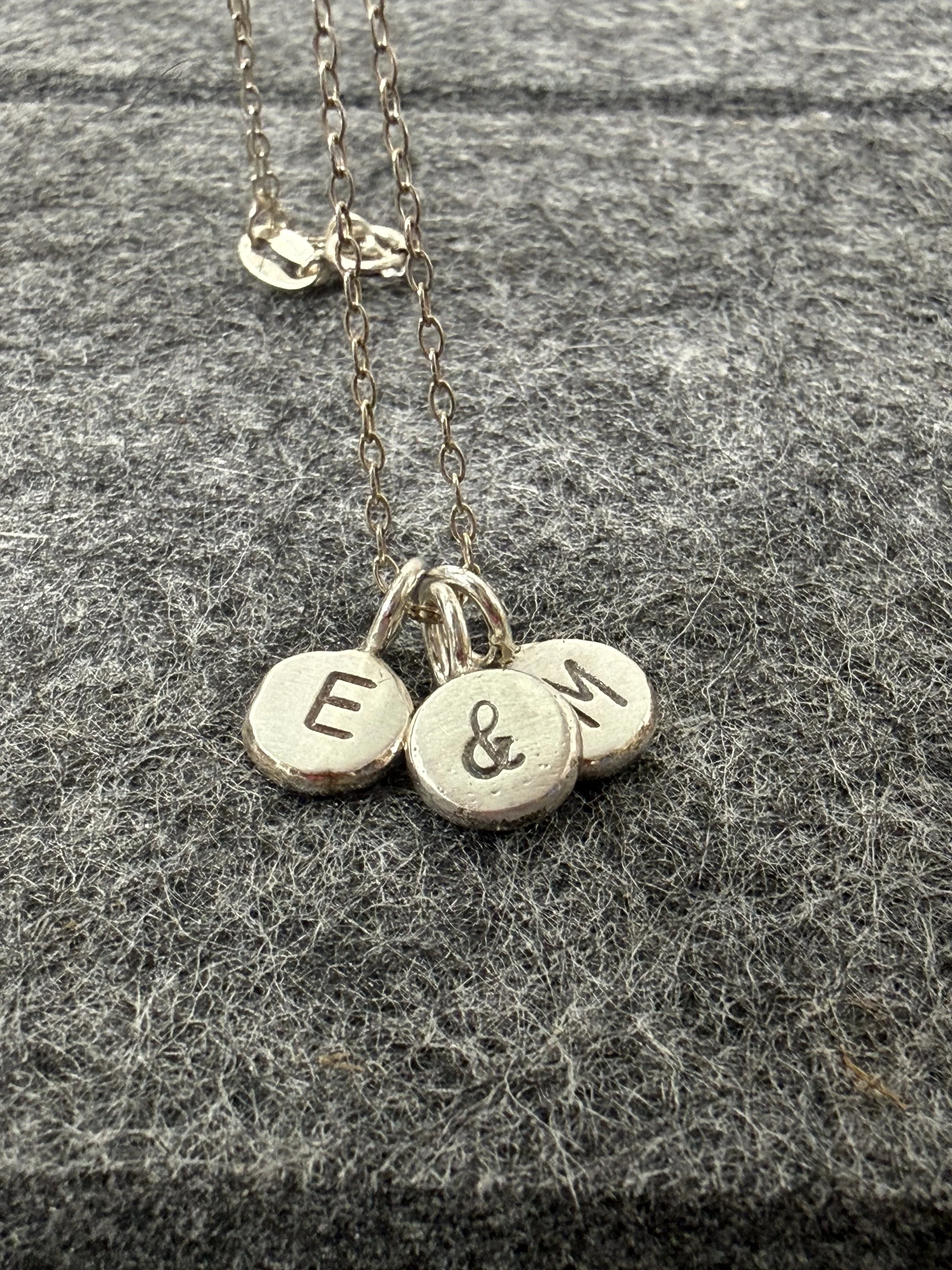 A picture of a delicate silver chain with 3 tiny pebble pendants with E&M stamped individually on each one.