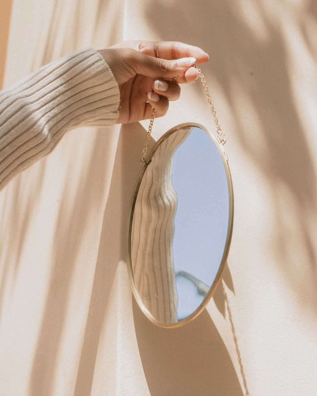 Hand hält einen runden Spiegel mit goldrand an einer goldkette, der an einer Wand hängt, die durch Sonnenlicht beleuchtet wird.