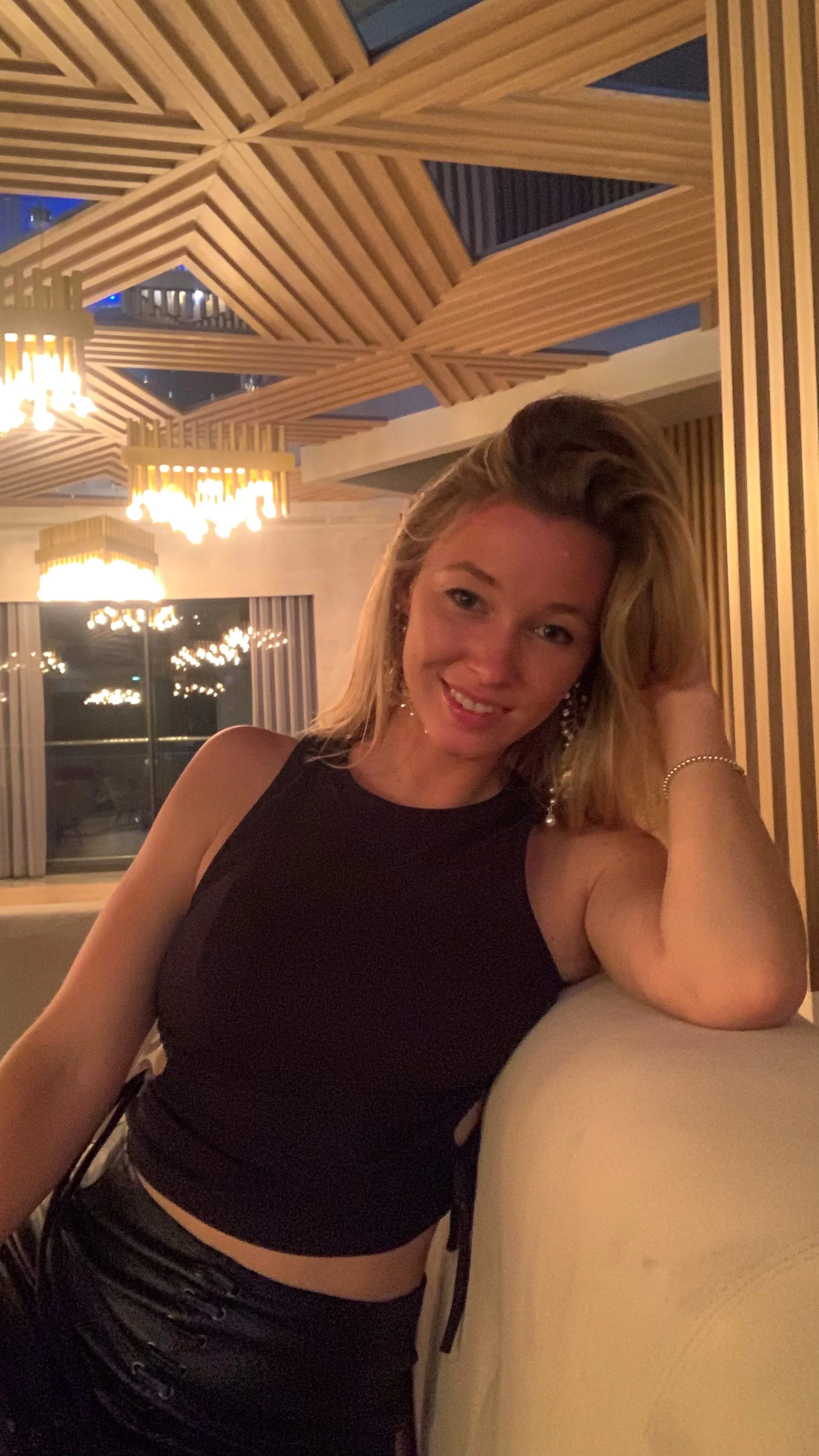 Junge Frau mit blonden Haaren in einem schwarzen Top sitzt in einem eleganten Raum mit geometrischen Holzdekor und Kronleuchtern.