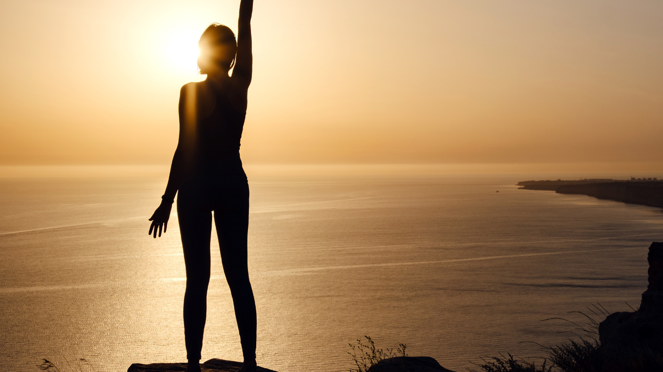 Eine silhouettierte Frau steht auf einer Klippe vor einem Sonnenuntergang über dem Meer, hebt einen Arm nach oben.