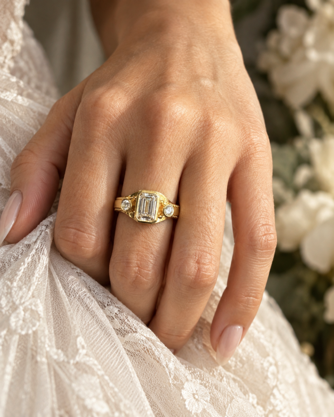 Handmade 18k gold emerald-cut diamond three-stone ring with round diamond side stones and fine granulation, worn on the hand.