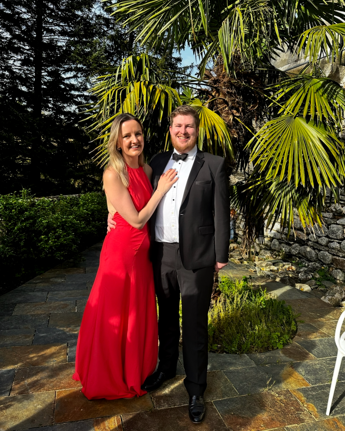 A man in a tuxedo and a woman in a red dress standing outdoors in front of green tropical plants, smiling for the camera.