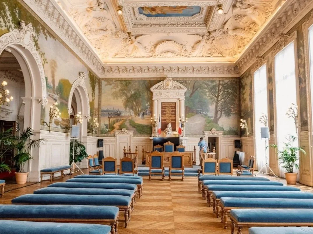 An elegant historical meeting room with blue upholstered benches and chairs arranged facing a raised platform with a judge's desk, a large ornate plaque, and decorative wall murals depicting a scenic landscape. The room features high ceilings with in