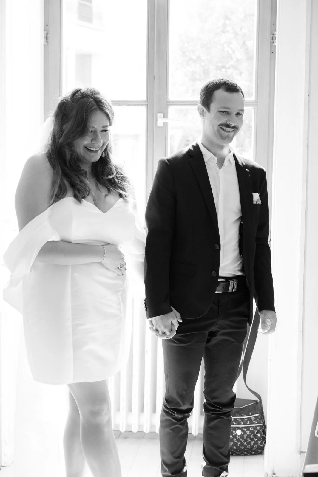 A bride and groom holding hands, smiling, standing indoors near a window.