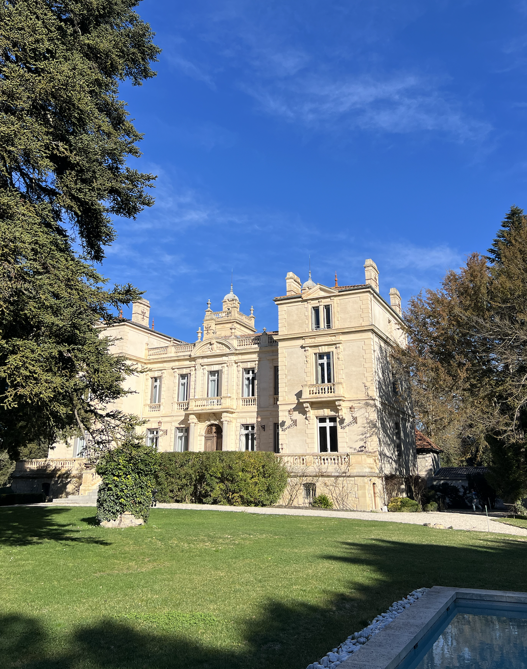 A large, elegant mansion with ornate architectural details, surrounded by trees and a well-maintained lawn, under a bright blue sky.