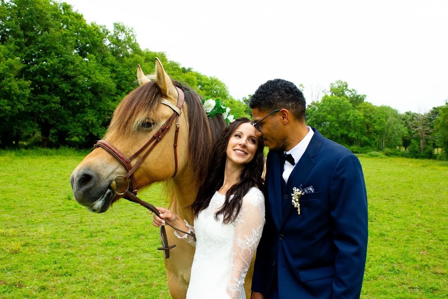 A bride and groom stand close together on a grassy field with a horse, smiling and looking at each other. The bride holds the horse's bridle.