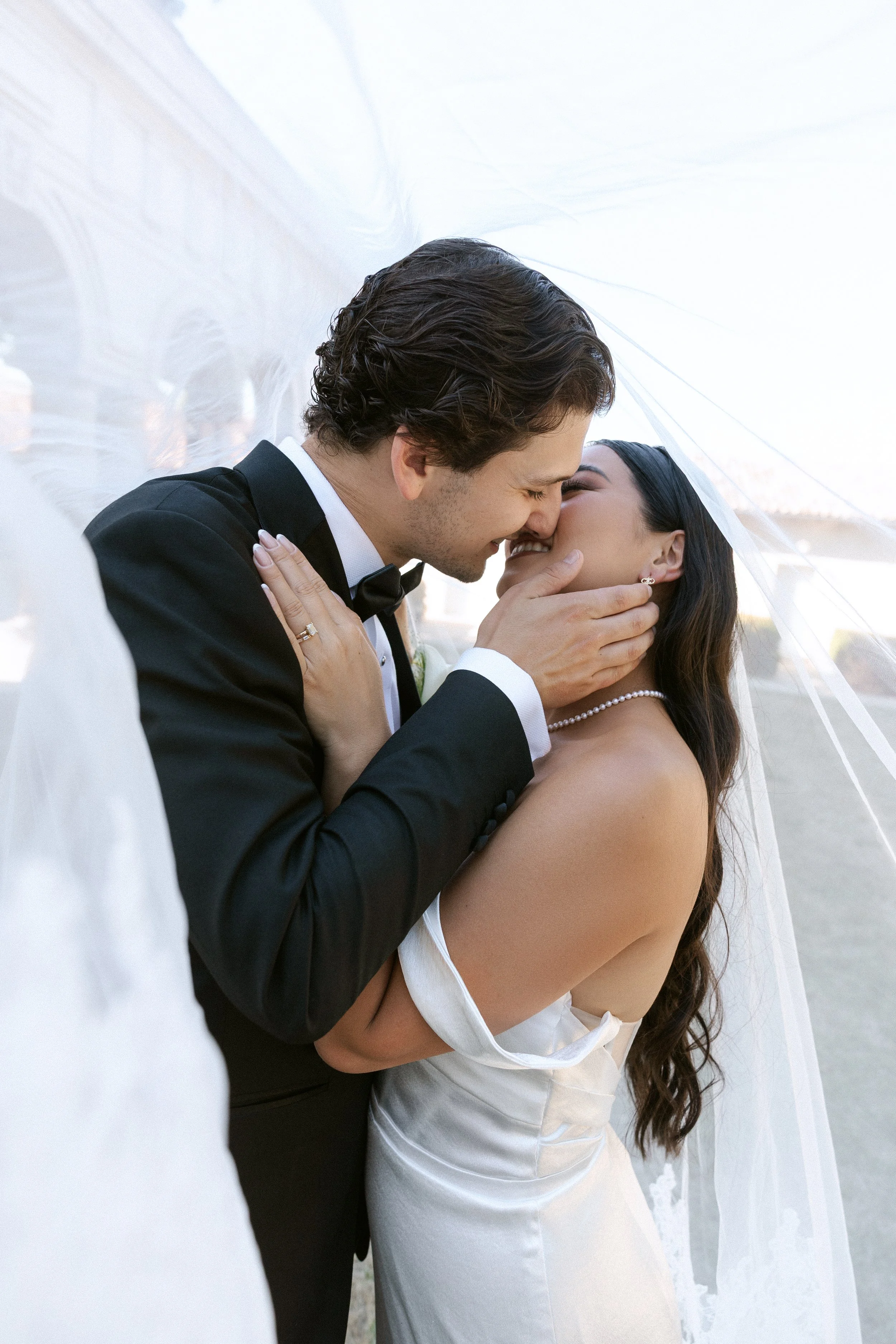 Wedding couple sharing a kiss under a sheer white veil, outdoors in natural light.