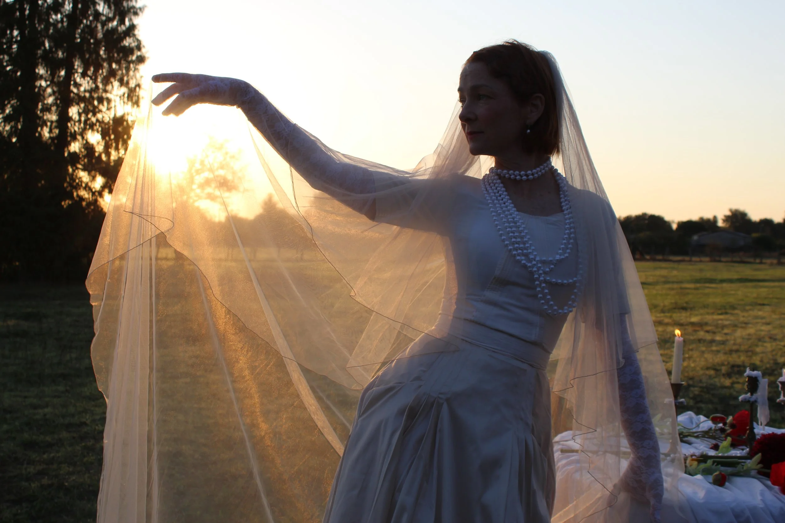 A woman dressed in vintage wedding attire stands outdoors at sunset, wearing a white dress, lace gloves, and multiple pearl necklaces, holding a sheer veil.