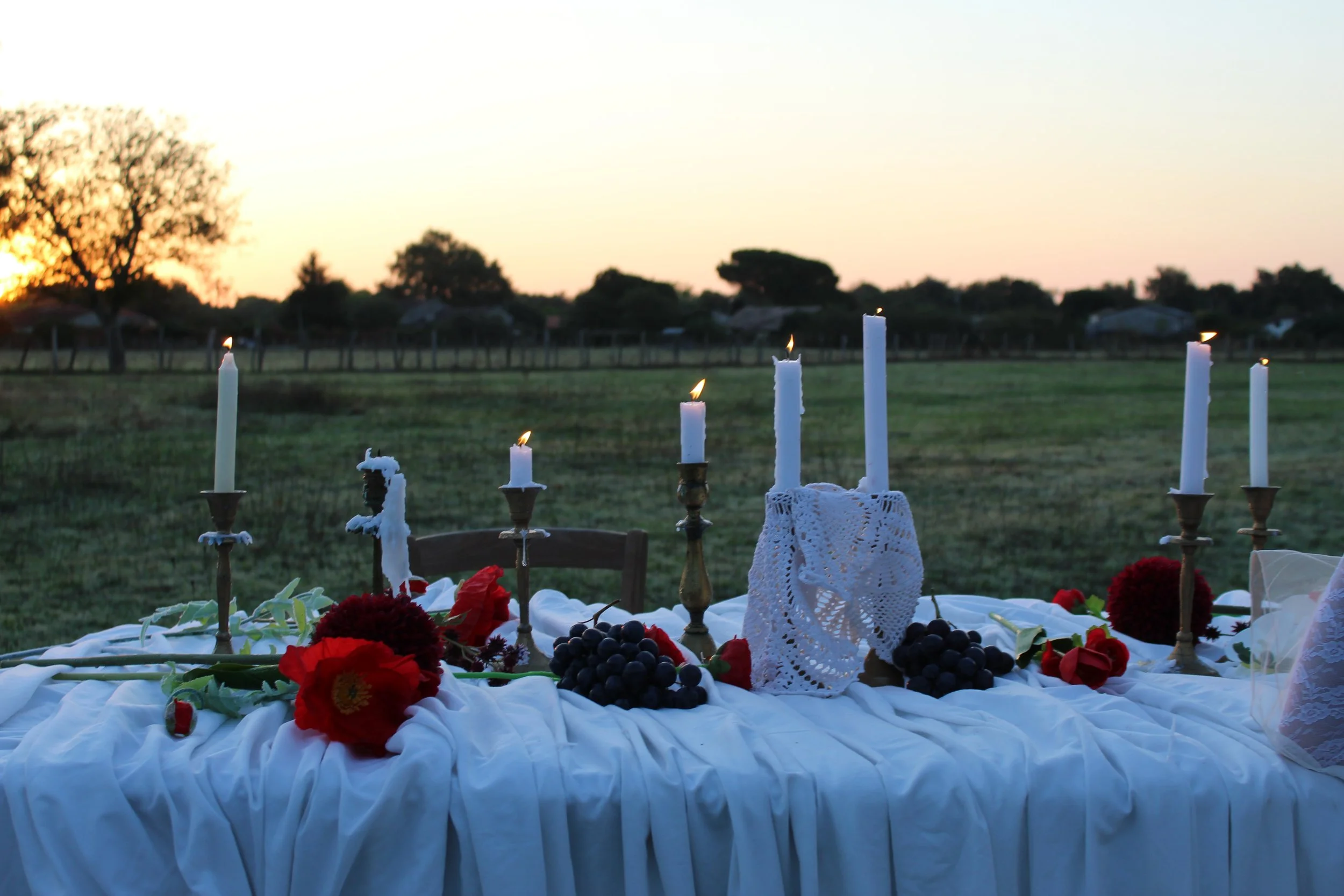 An outdoor table decorated with white cloth, red flowers, grapes, and lit white candles at sunset.