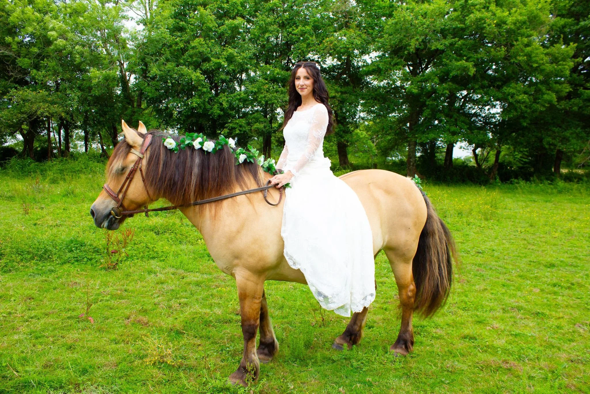 A woman in a white bridal dress riding a light brown horse with a floral headband on its mane, in a green outdoor setting with trees in the background.