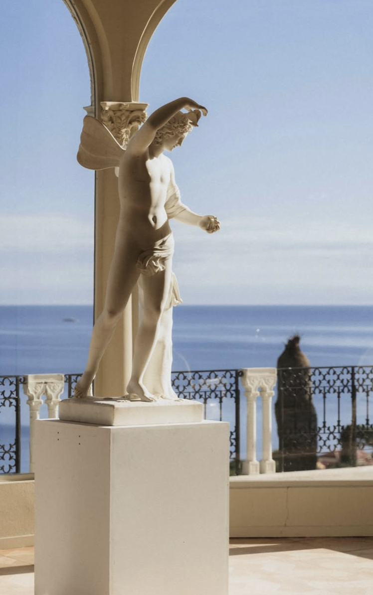 A white marble statue of a young man with a cloth draped around his waist, standing on a pedestal on a balcony with an ocean view. The statue's arm is raised, and a cat is sitting outside by the stone railing.